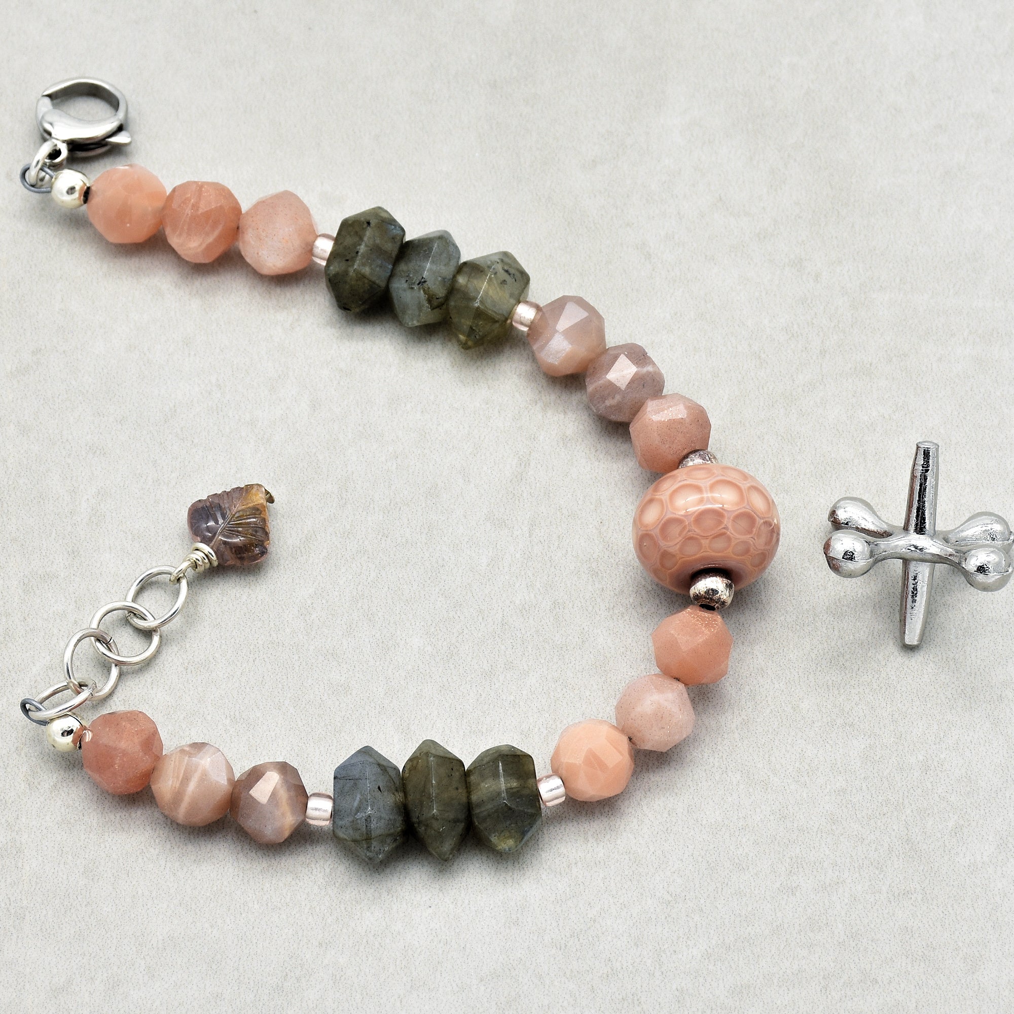 This image shows a bracelet. It is made of a variety of faceted beads in pink, peach, and greenish-gray tones, with a silver toggle clasp visible on the right side. The size and type of clasp, along with the length and bead arrangement, indicate that it is a bracelet.