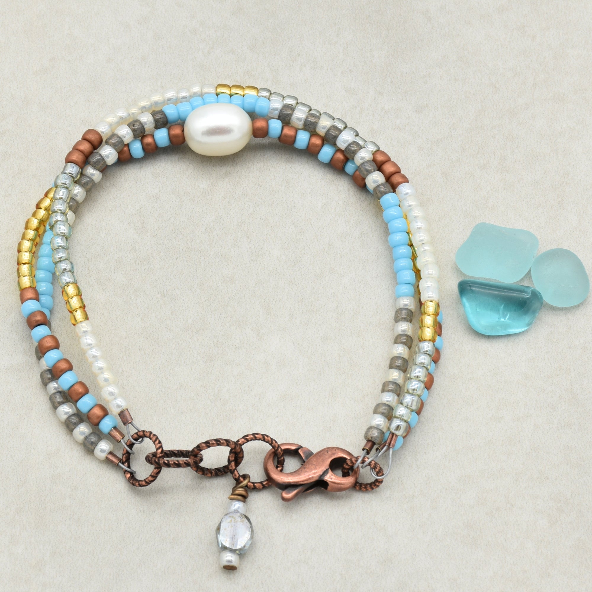 The image shows a bracelet. It is a beaded multi-strand bracelet with small beads in various colors (blue, gold, silver, brown, and white) and a larger white bead as a focal point. The bracelet has a copper clasp and a small dangling bead charm, which are typical features of bracelets.