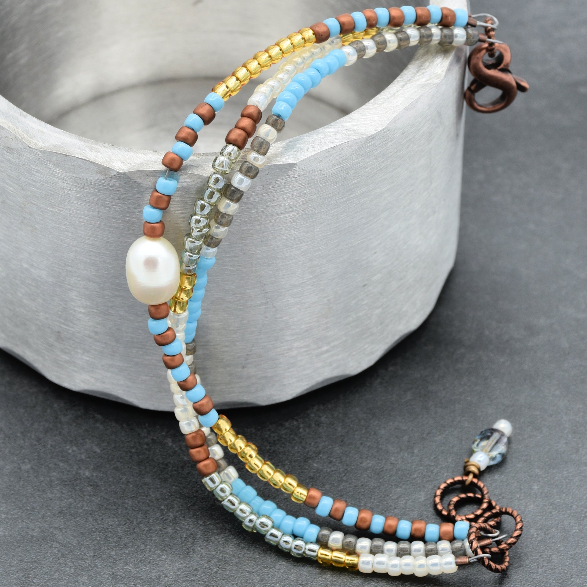 This image shows a bracelet. It consists of multiple strands of small beads in blue, copper, gold, silver, and clear colors, along with a single white pearl bead. The bracelet has a clasp at one end, confirming that it is designed to be worn around the wrist.