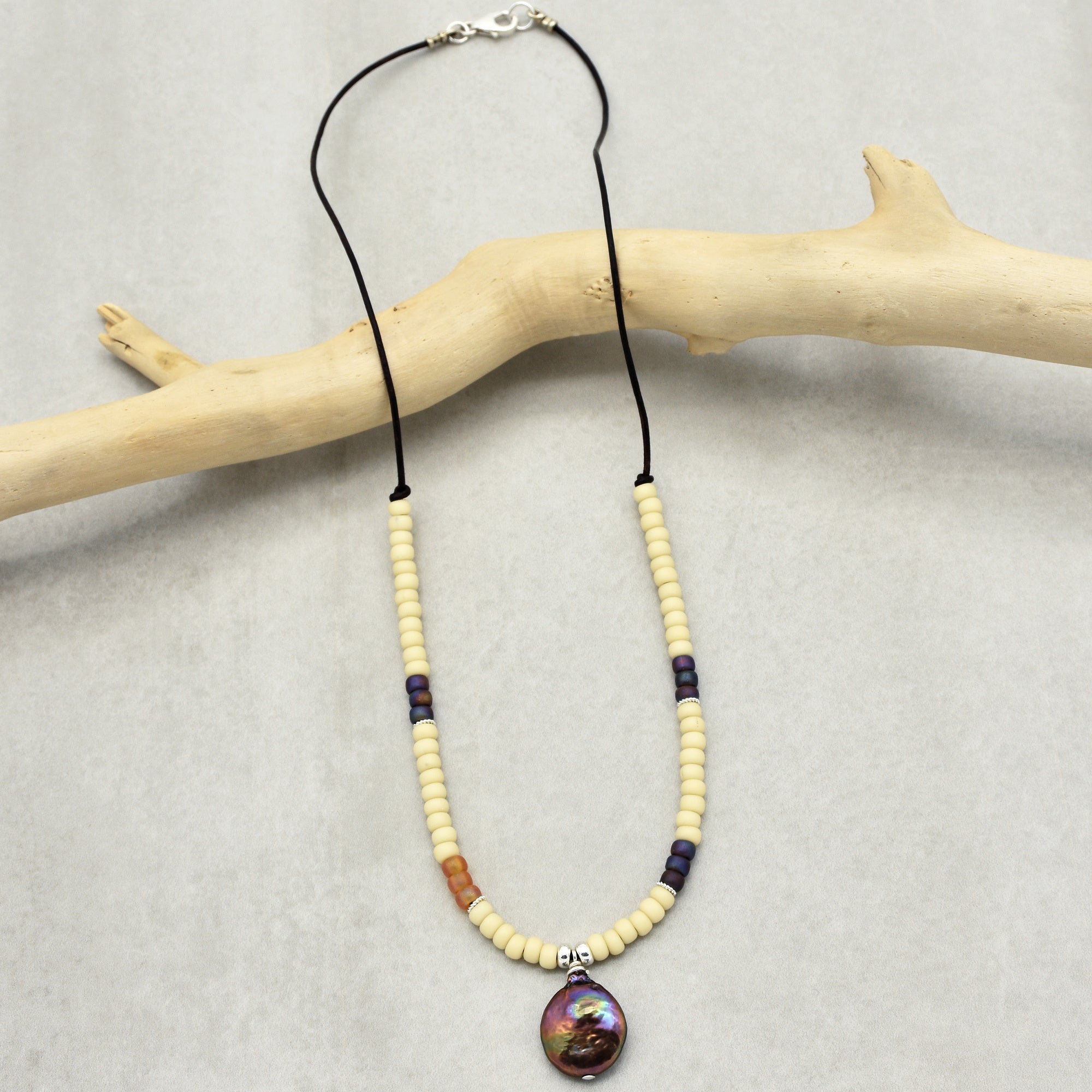 This image shows a necklace. It has a strand of beads and a larger, central pendant, and it is displayed on a light surface with a piece of driftwood as a prop.