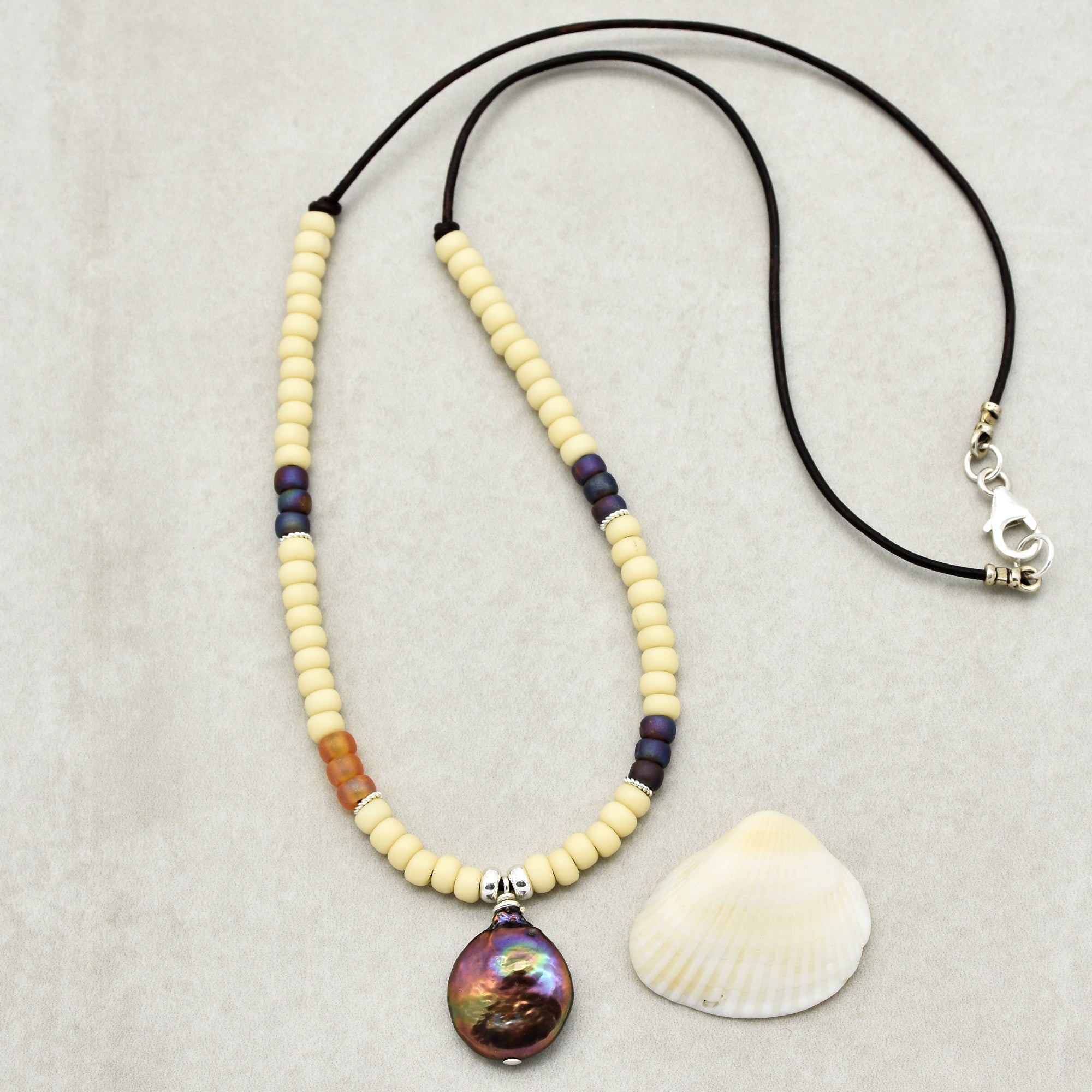 This image shows a necklace. It features a string of cream-colored and dark beads with a central iridescent shell pendant, and it is attached to a black cord with a clasp for fastening around the neck.