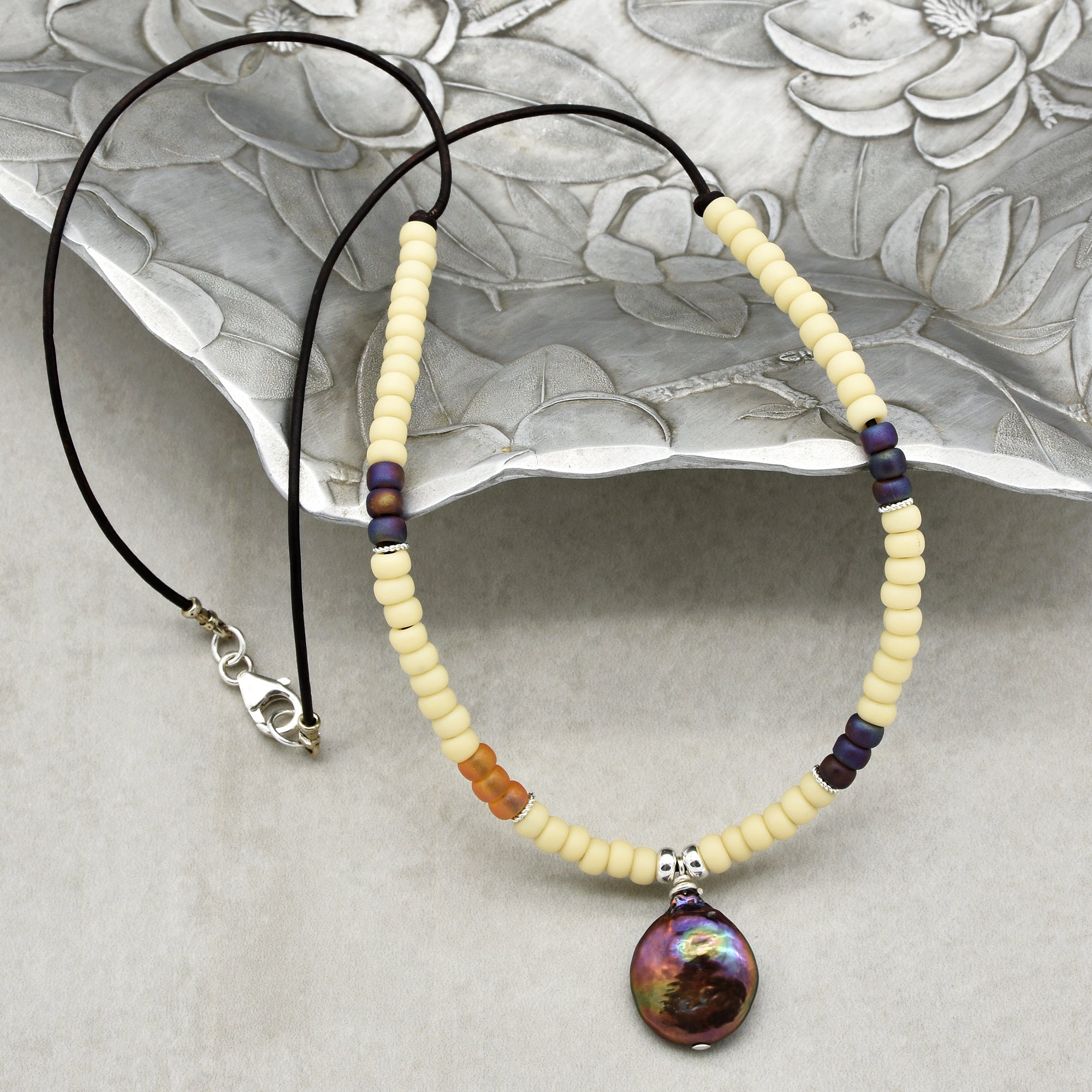 This image shows a necklace. It features a combination of cream-colored and purple beads strung on a cord, with a large, iridescent pendant hanging in the center. There is also a clasp visible on one end, which is typical for necklaces.