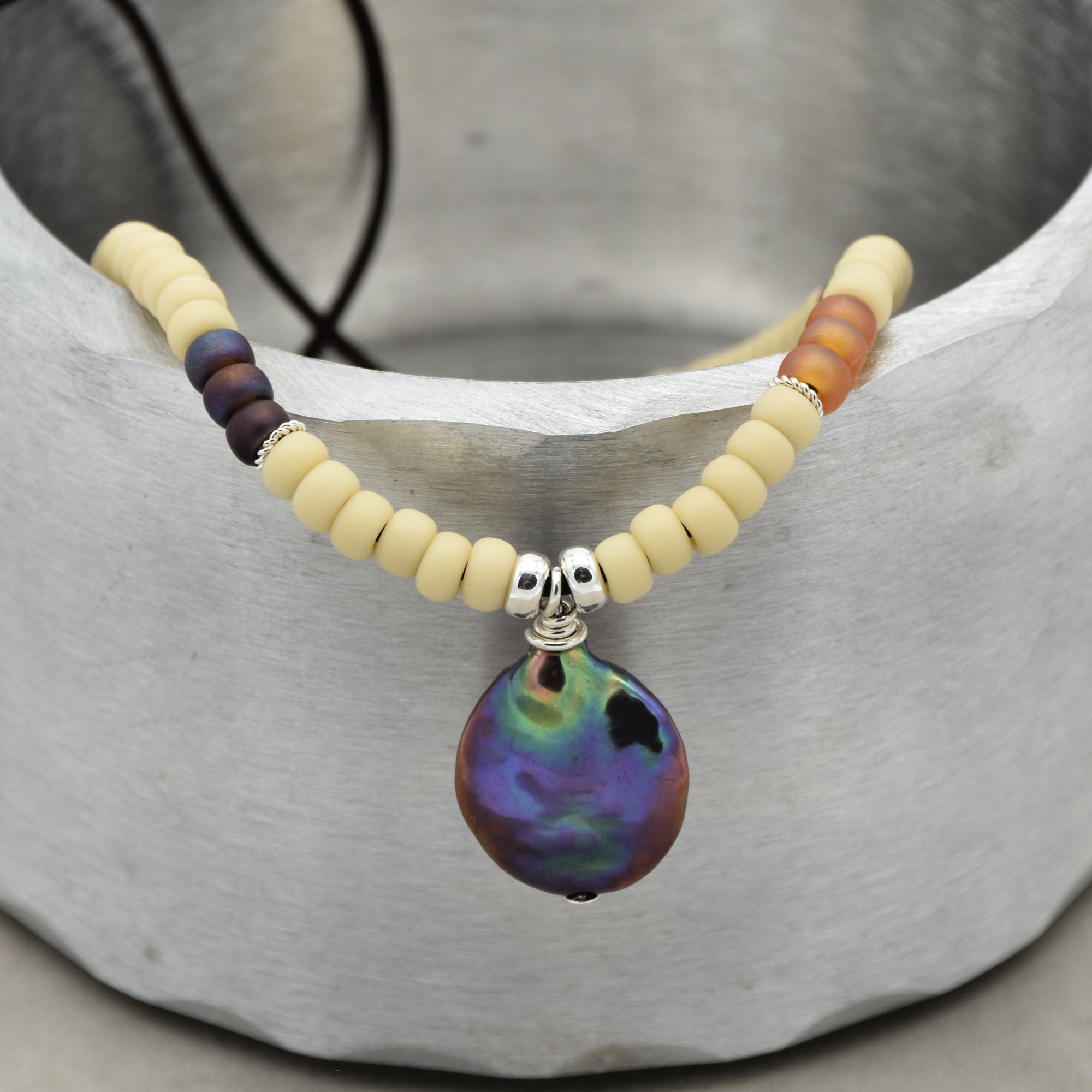 The image shows a necklace. It has cream-colored and some orange and brown beads with a large, iridescent pendant hanging from the center.