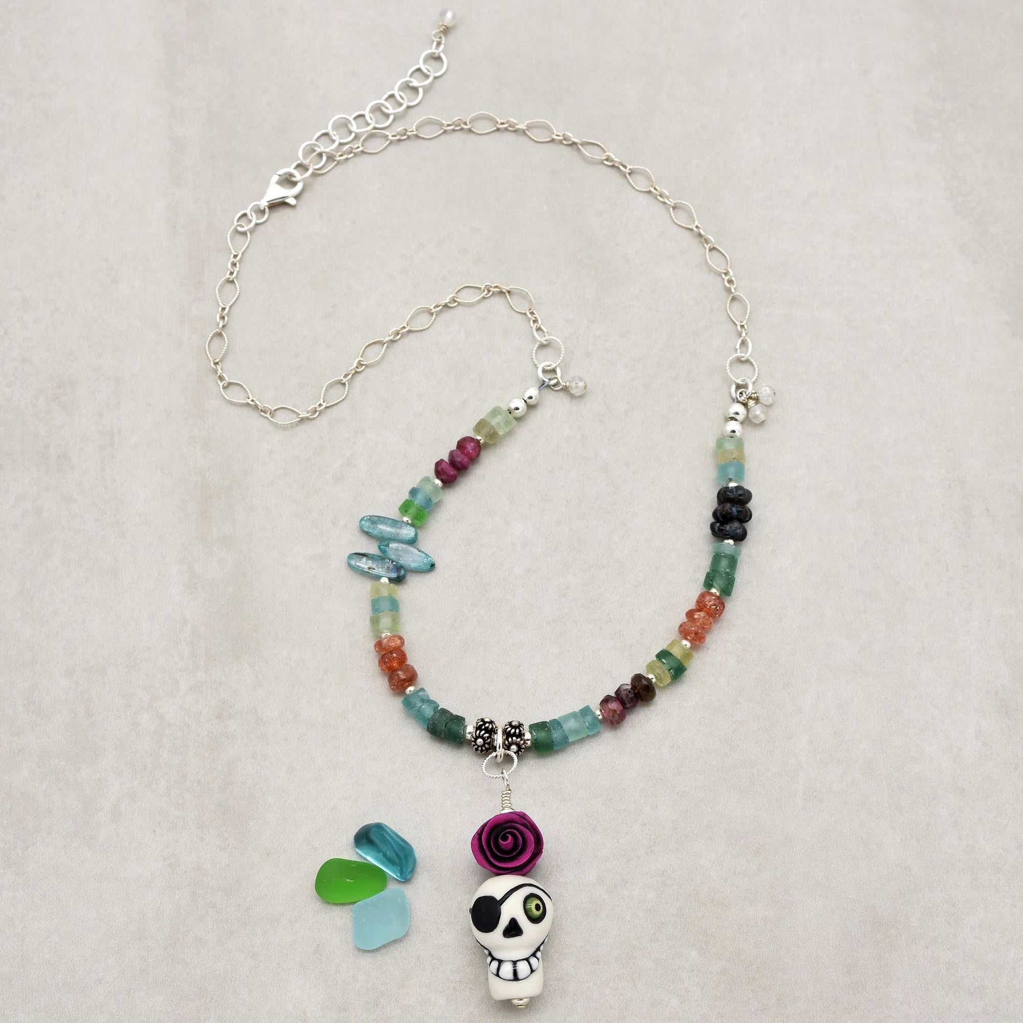This image shows a necklace. The piece of jewelry has a chain with various colorful beads and a skull pendant with a pink rose on top. The length and the design, including the chain and clasp, indicate that it is intended to be worn around the neck.