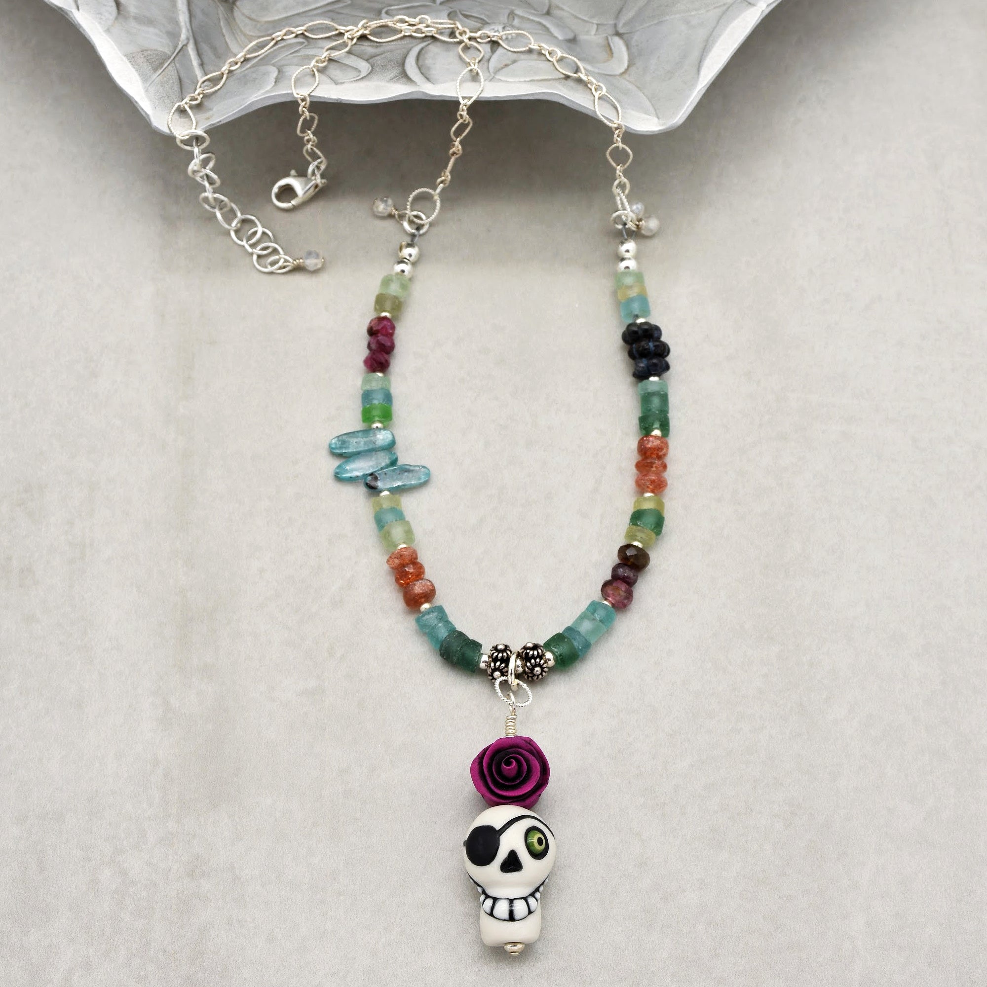 This image shows a necklace. It is composed of colorful beads and features a pendant with a skull wearing an eye patch and a purple rose above it. The necklace also has a silver chain and clasp.