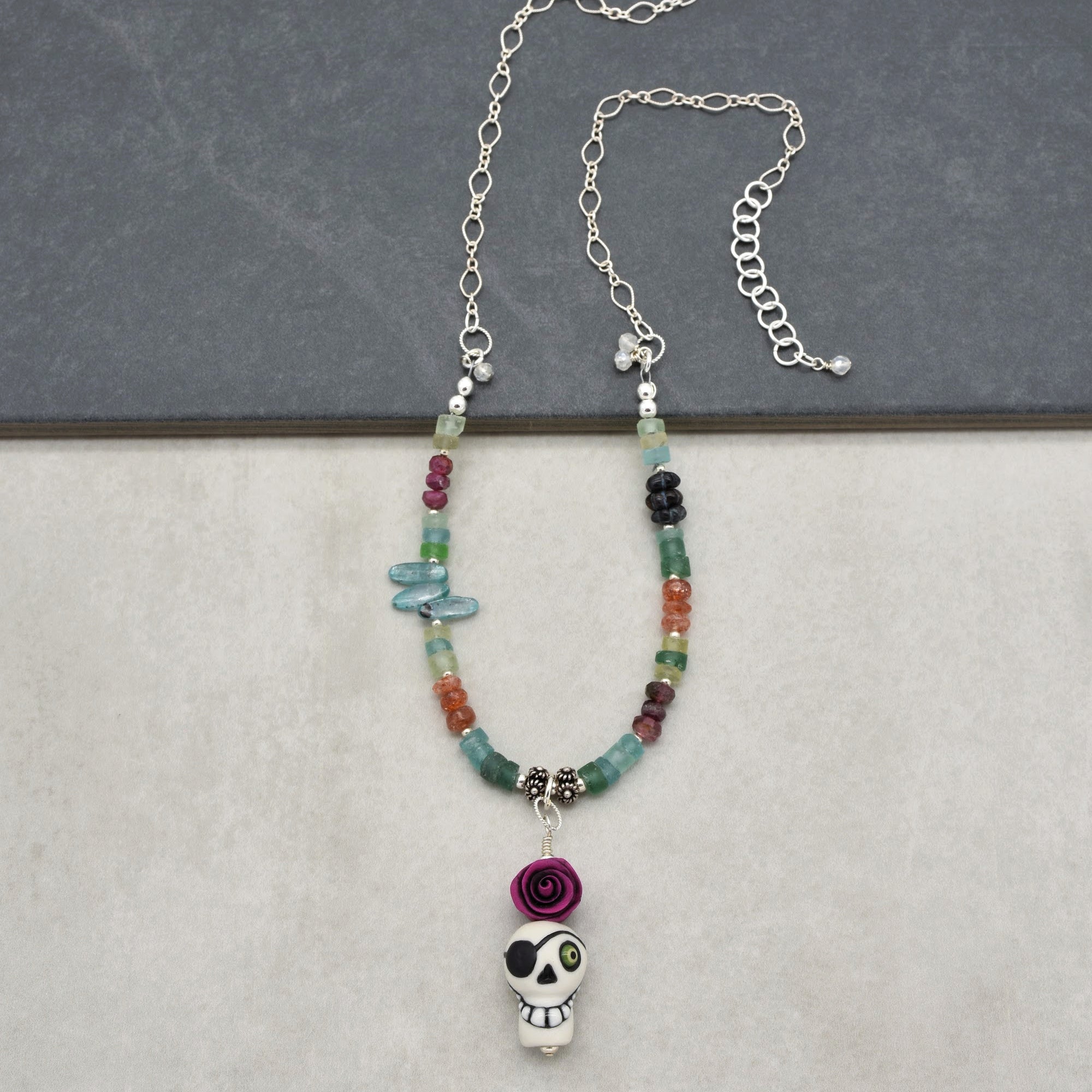 This image shows a necklace. It has a chain with an adjustable clasp and a series of colorful beads. The necklace features a pendant with a skull design wearing an eye patch and decorated with a purple rose above it.