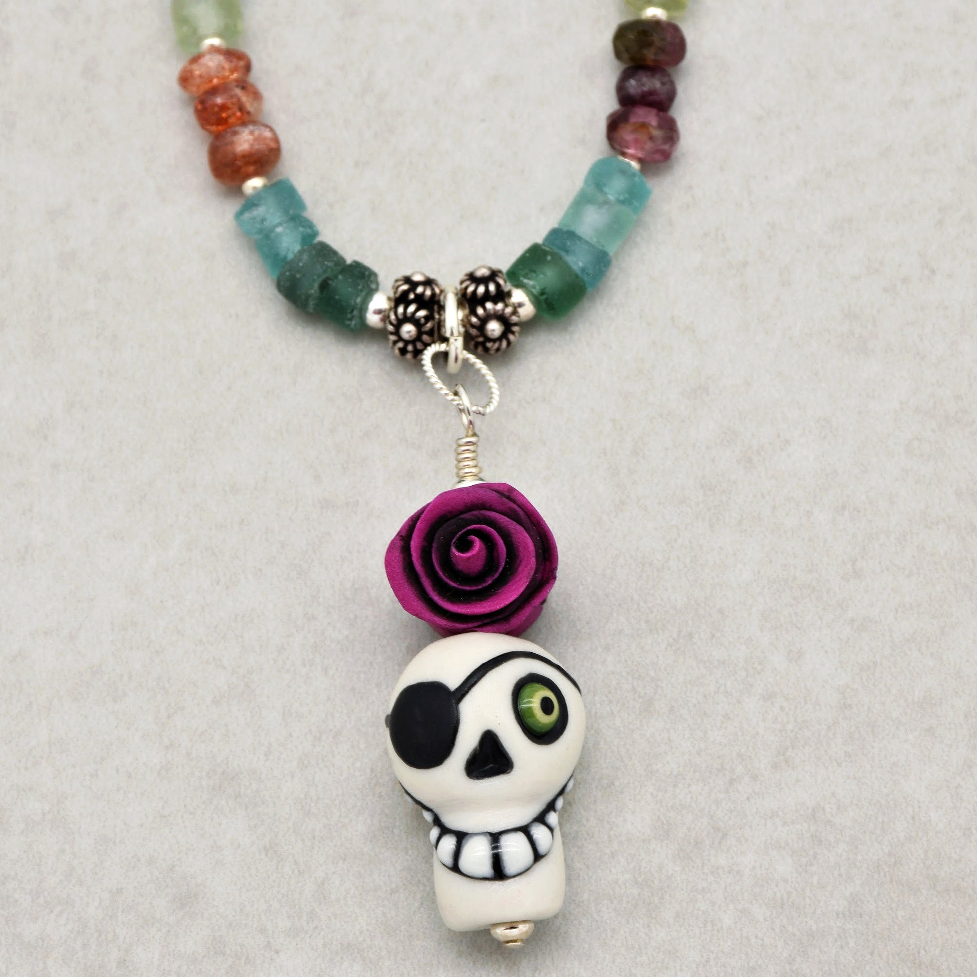 This image shows a necklace. The jewelry piece features a pendant with a skull wearing an eyepatch and a purple rose above the skull, attached to a beaded string.