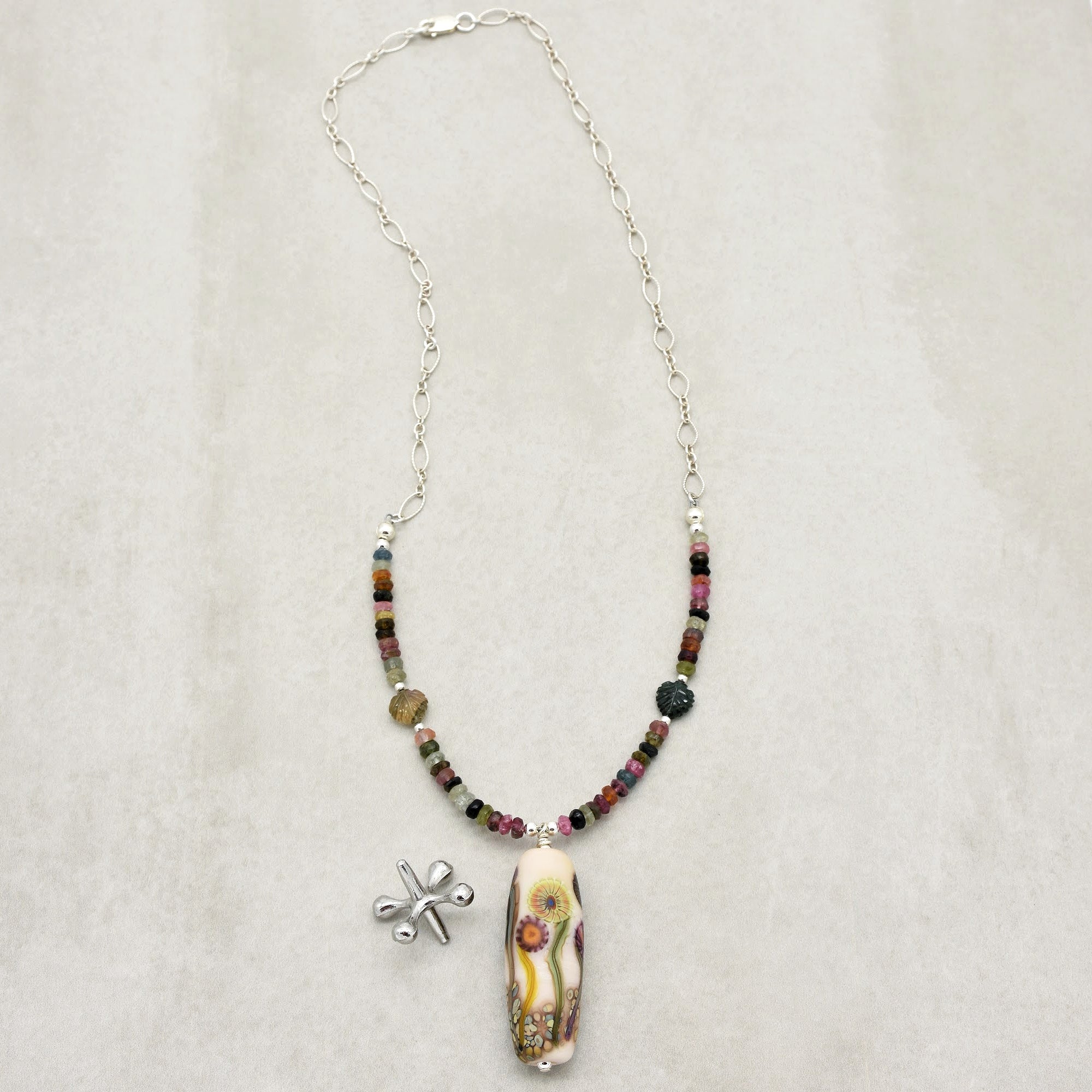 The image shows a necklace. It has a chain with colored beads and a decorative pendant featuring a floral design. There is also a small silver charm or accessory next to it.