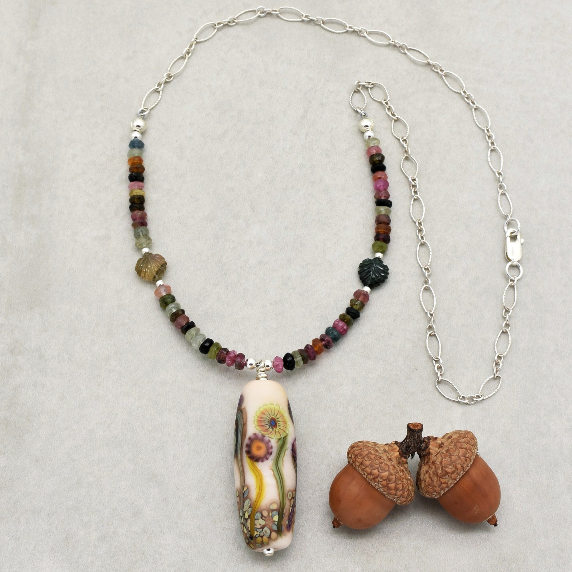 This image shows a necklace. It has a silver chain with multicolored beads and leaf-shaped beads, along with a central elongated pendant featuring a floral design.