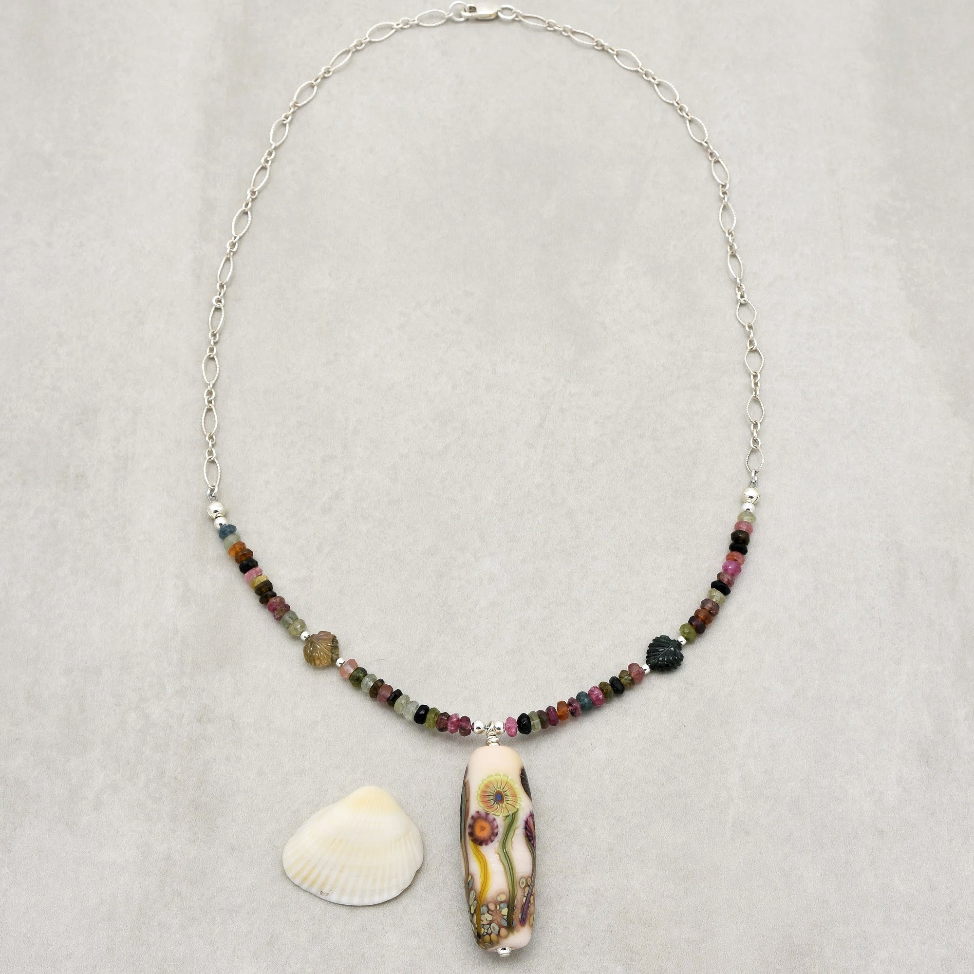 This image shows a necklace. The chain is made of linked silver loops, and it features colorful beads along the front. There is a central large pendant with a floral design. The overall shape and length of the chain indicate that it is a necklace rather than an earring or bracelet.