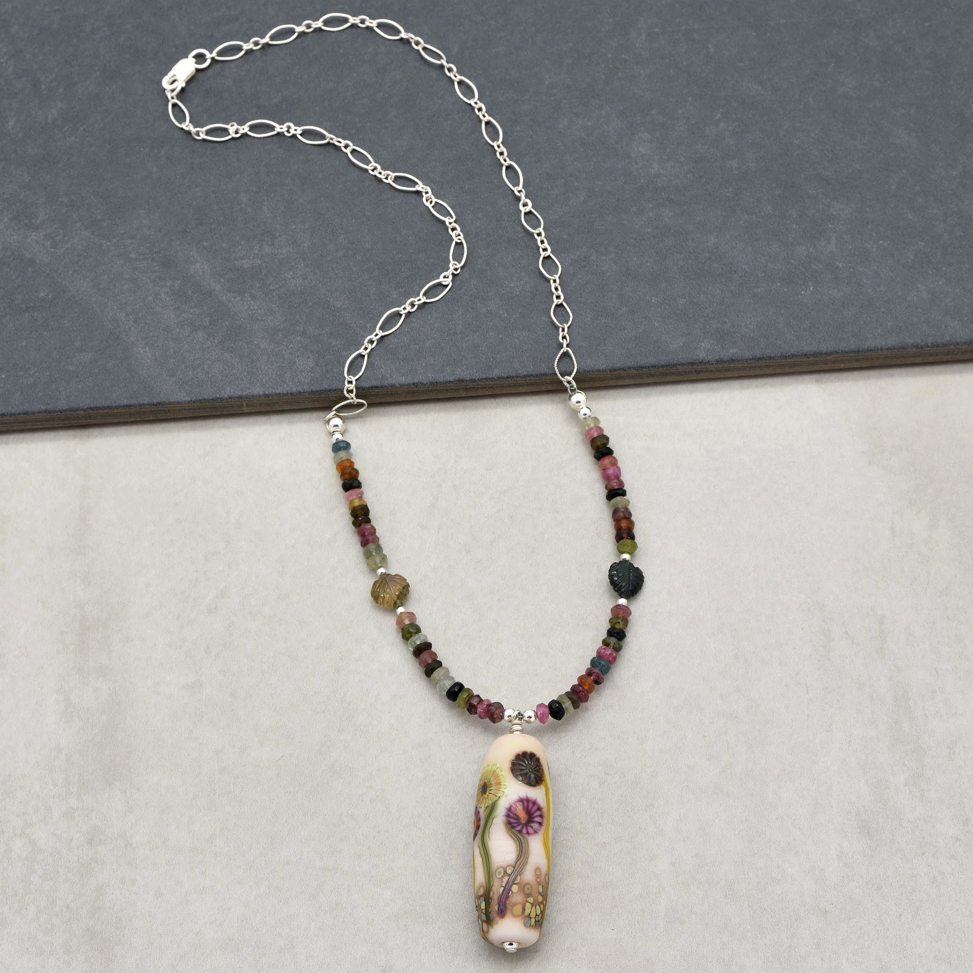 This image shows a necklace. It features a thin, silver chain with a series of small, colorful beads near the center, and a decorative pendant with floral designs hanging at the bottom.