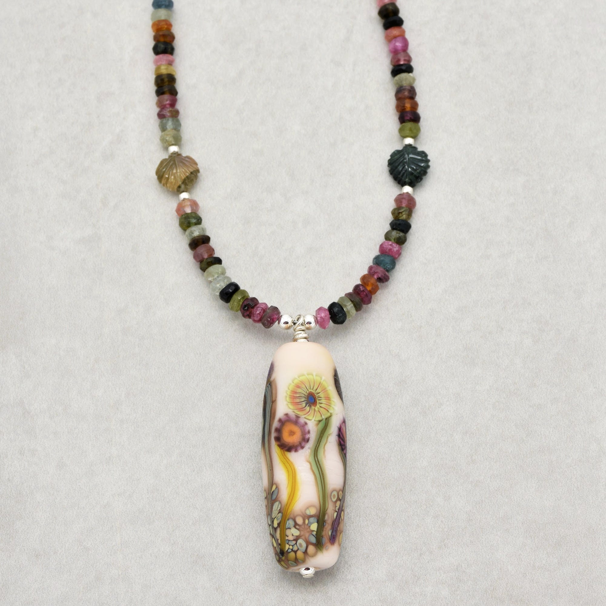 This image shows a necklace. It features a string of multicolored beads and a central pendant with a floral design.