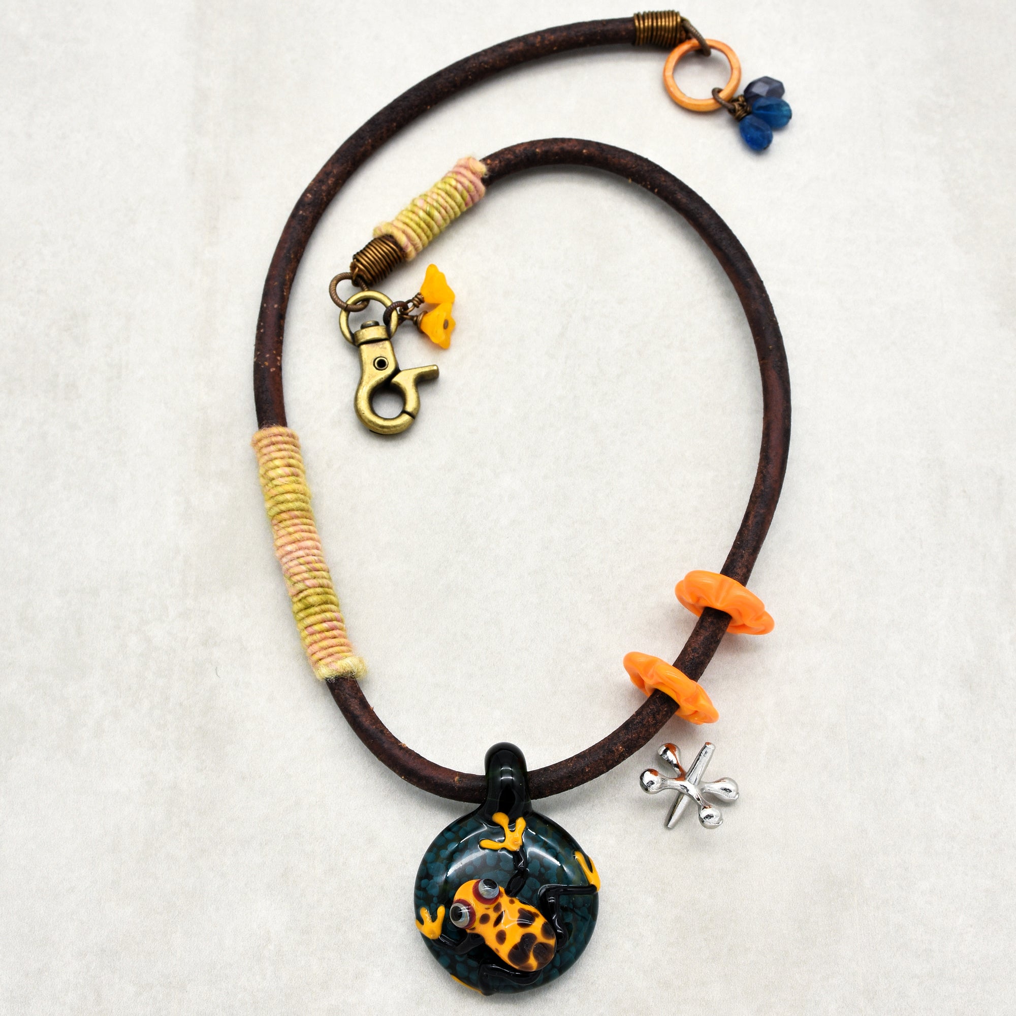 This image shows a necklace. It has a circular pendant with a frog design, attached to a thick cord with decorative orange rings, wrapped thread sections, and a clasp.