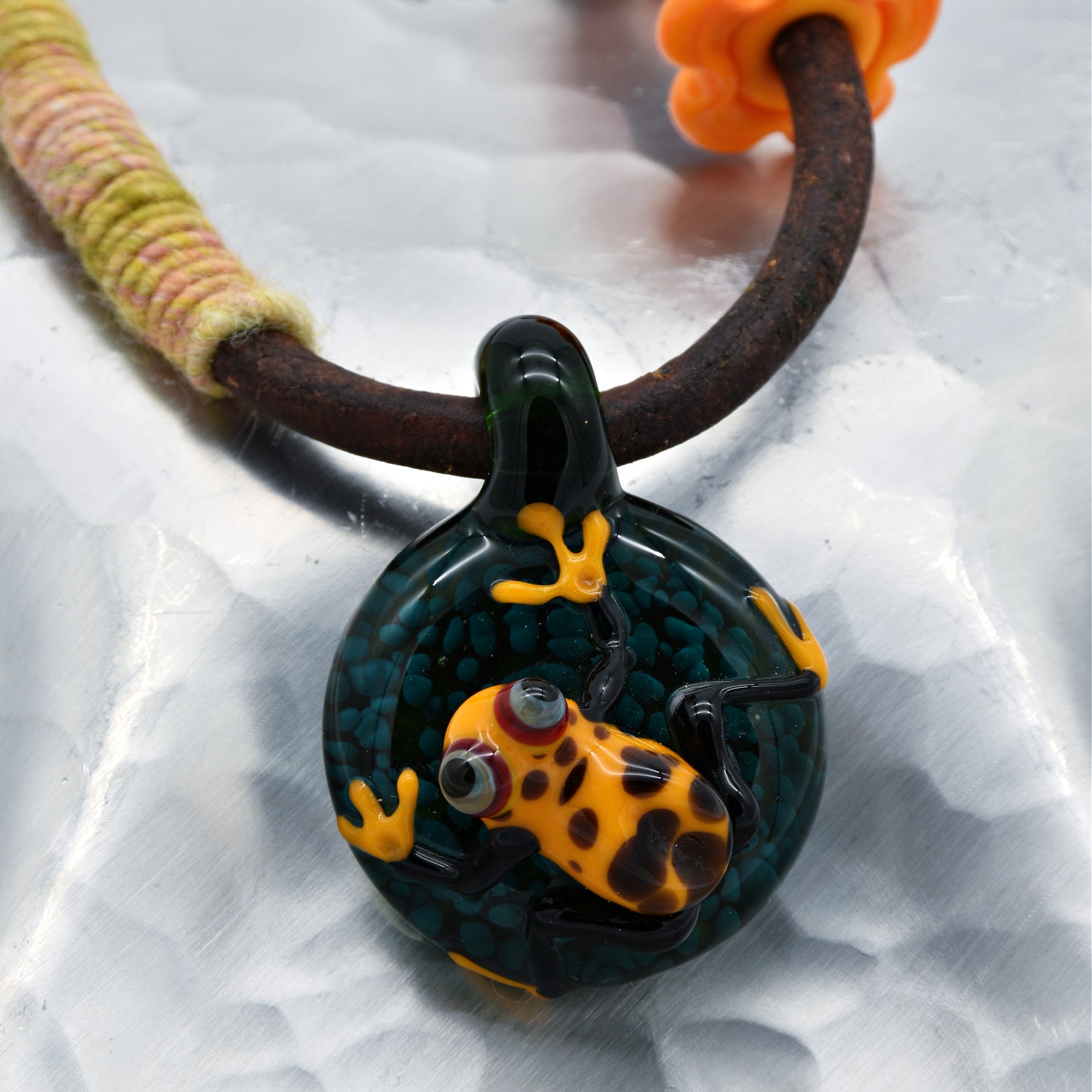 This image shows a necklace. The jewelry piece is characterized by a leather or rustic cord with a pendant. The pendant has a detailed glass design featuring an orange and black spotted frog. The cord is partially wrapped with colorful thread near the edges.