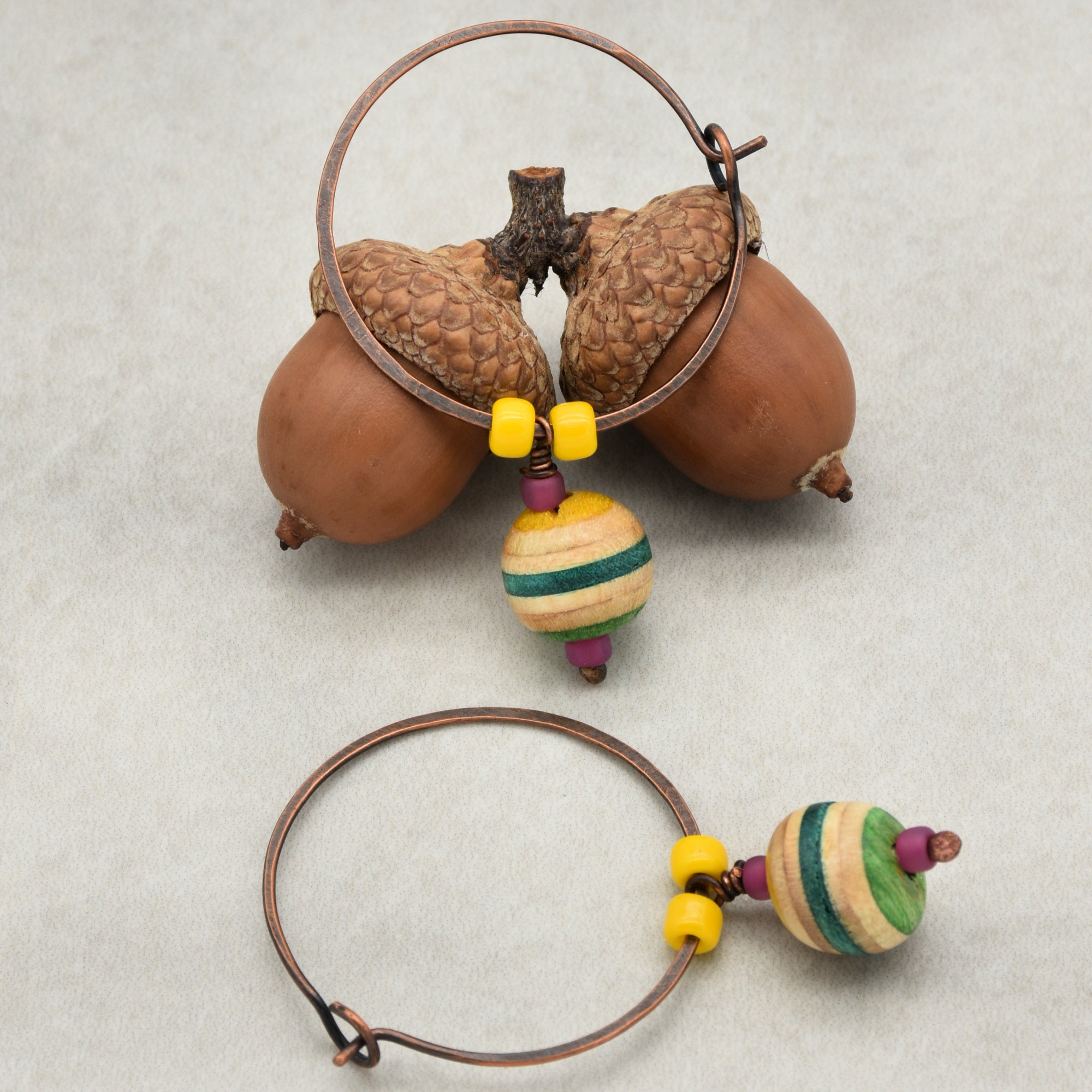 The image shows a pair of earrings. They are circular hoops made of metal with decorative beads attached, including a colorful round wooden bead and two small yellow beads on each earring. The earrings are displayed with two acorns.