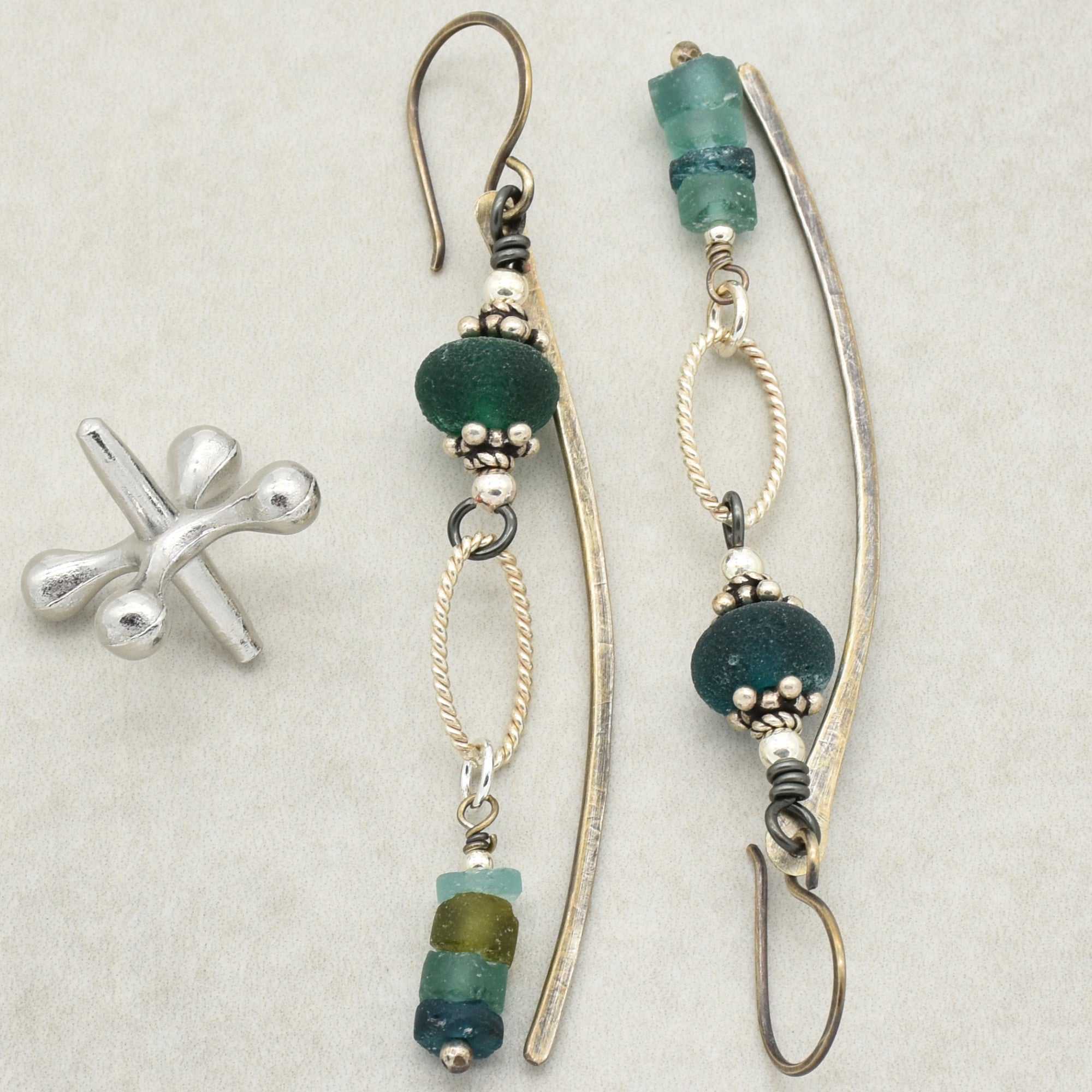 The jewelry shown in the image are earrings. They feature elongated, slightly curved metal pieces with decorative beads in shades of green and blue attached, along with twisted oval-shaped metal links. There is also a small, separate silver cuff or ear climber piece visible in the image.