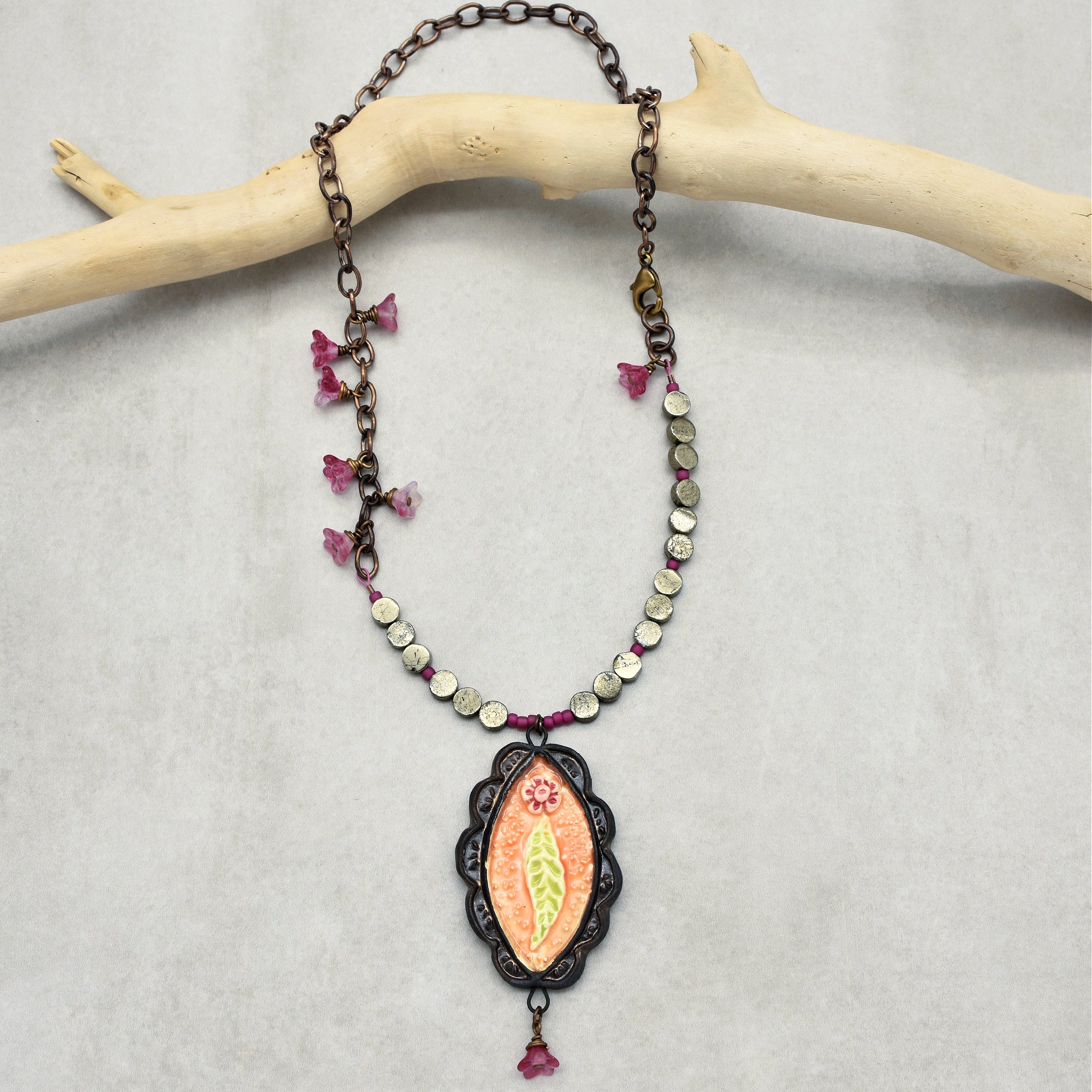 This image shows a necklace. It features a chain with decorative beads and a central pendant with a floral design. The necklace is displayed hanging on a piece of light-colored wood.