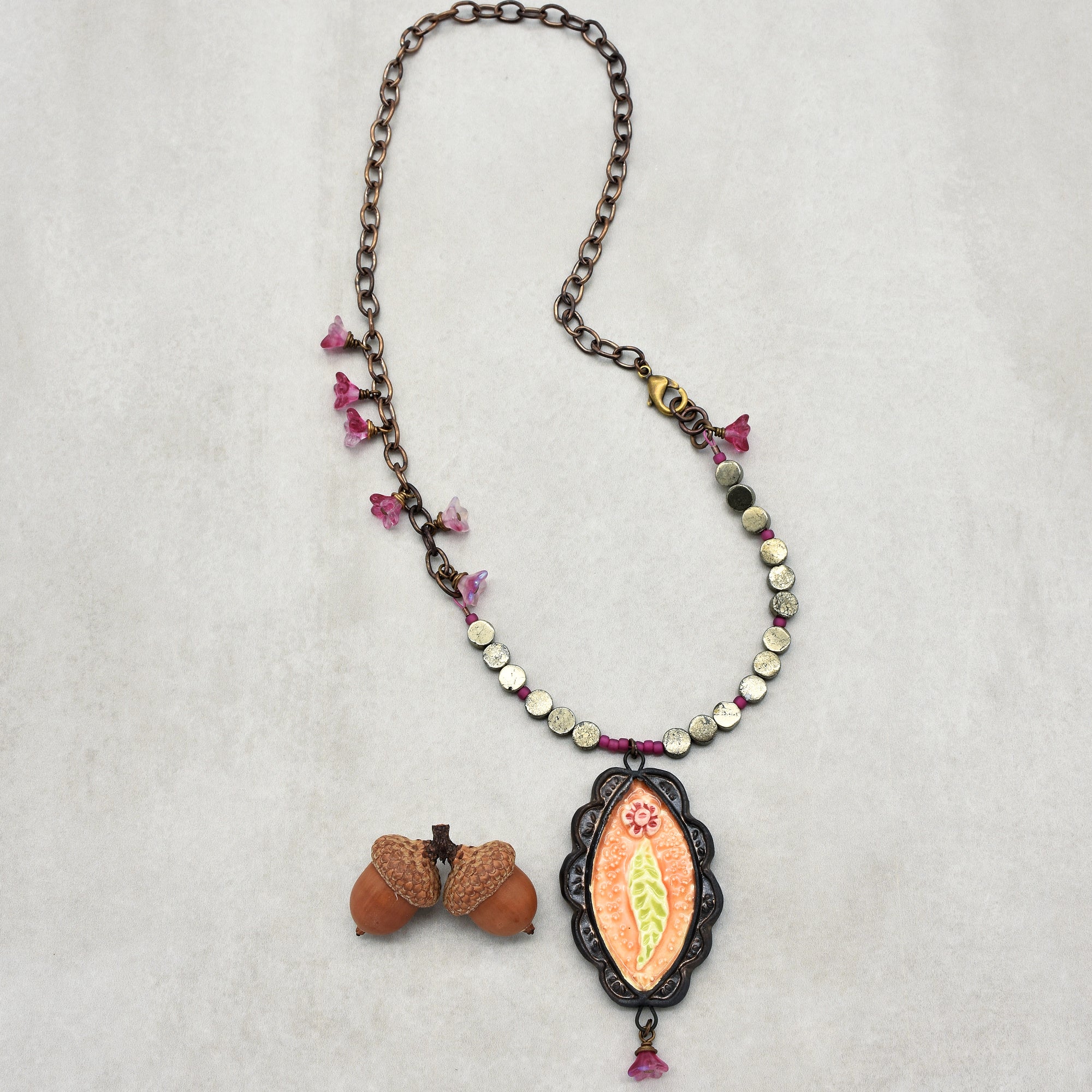 This image shows a necklace. It has a chain with small pink flower-shaped decorations, round metallic beads, and a central pendant with a floral design. There is also a pair of acorns placed next to the necklace.