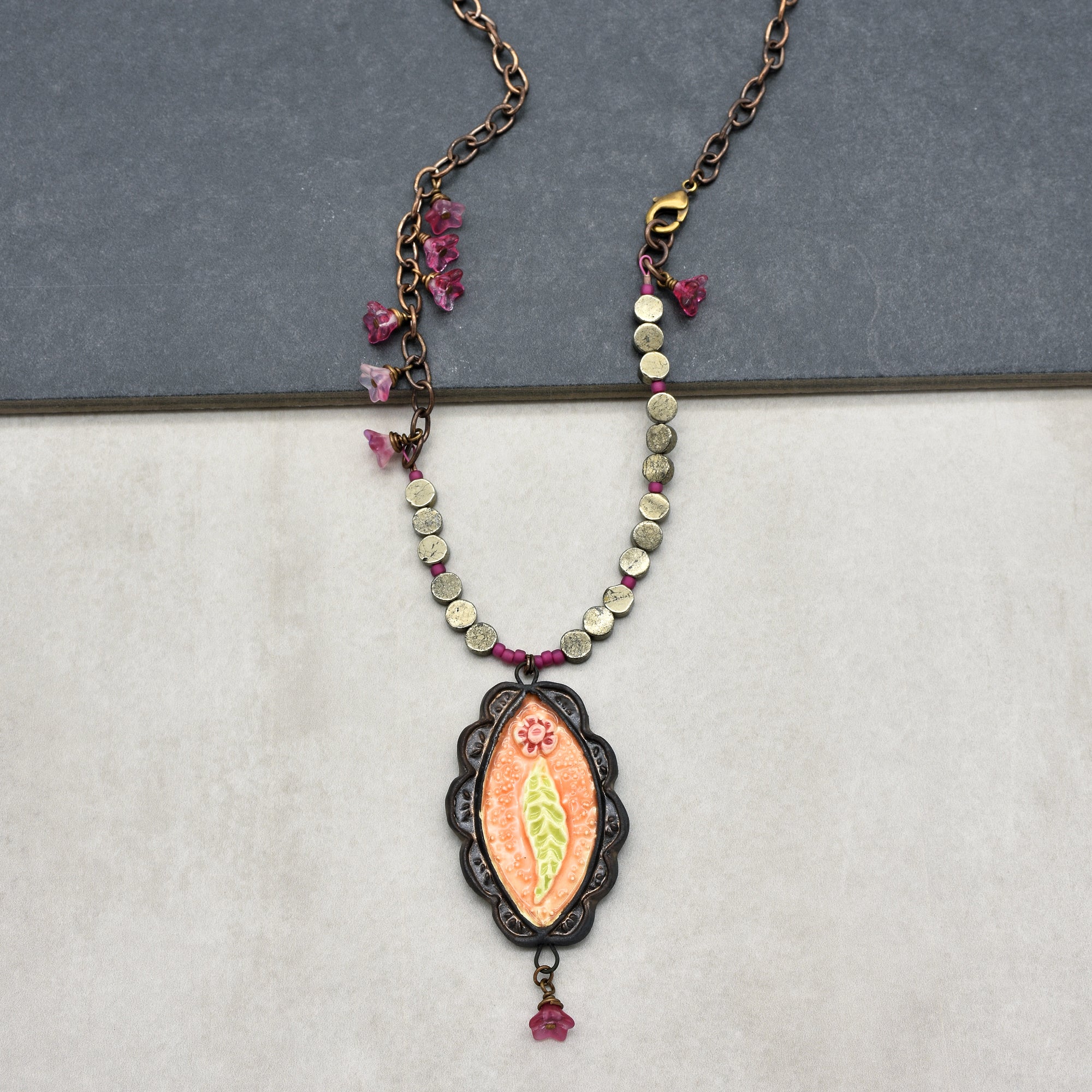 This image shows a necklace. It has a chain with decorative beads and a central pendant featuring a floral design and a scalloped black frame.