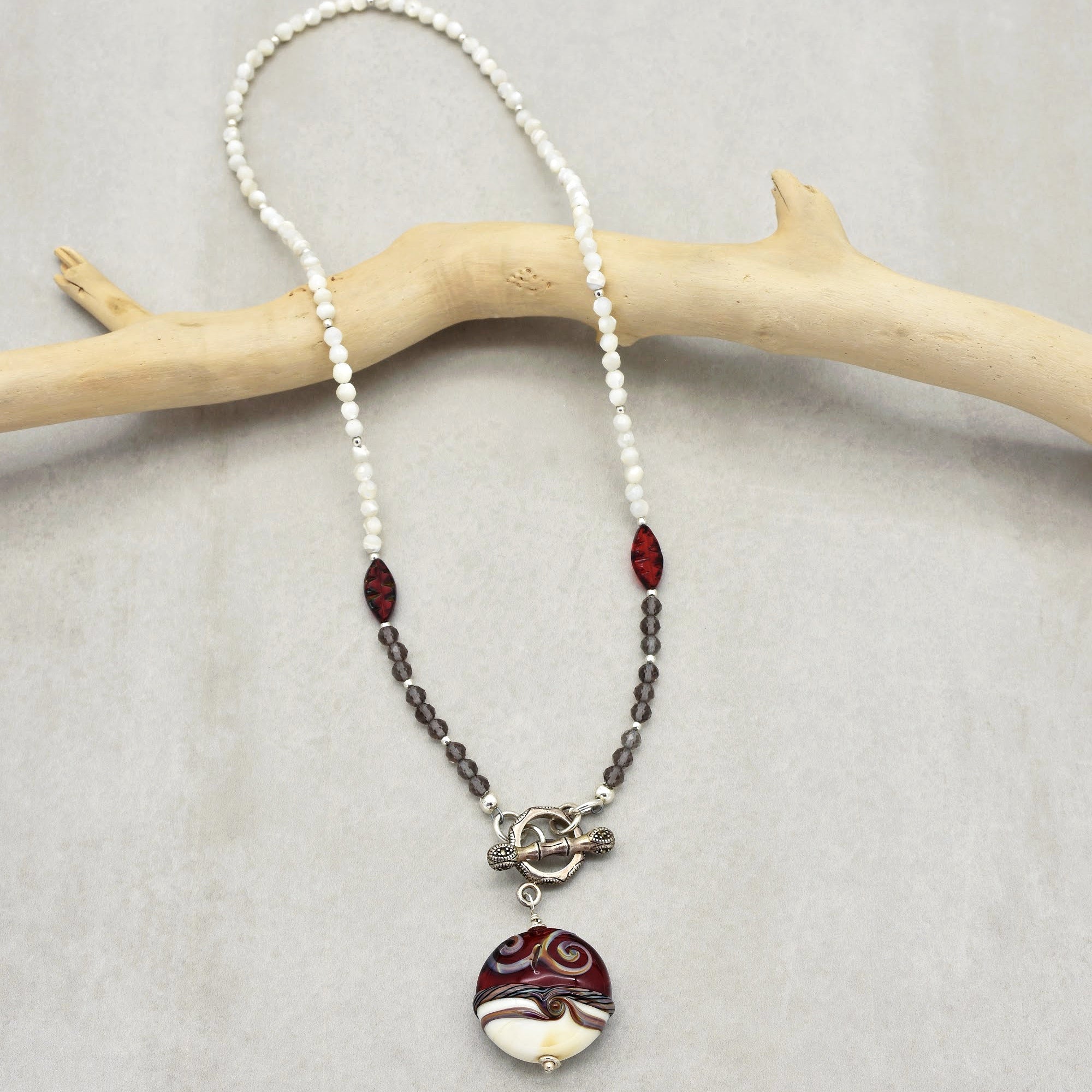 This image shows a necklace. It features a beaded design with small white and darker beads, along with two larger red beads. The necklace has a toggle clasp and a prominent pendant with a swirl pattern in red, white, and brown colors.
