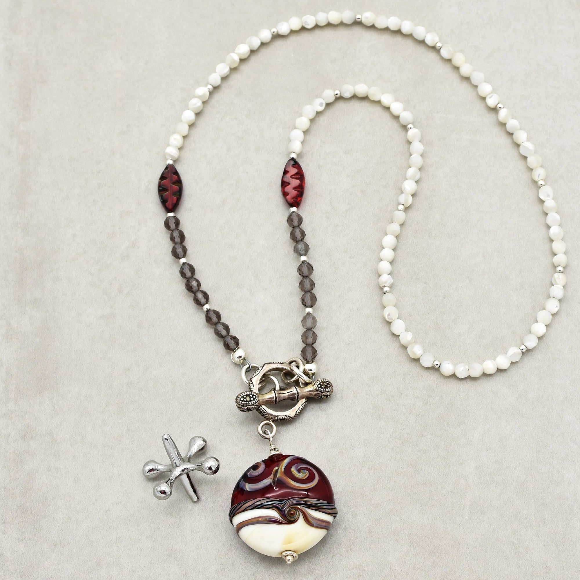 This image shows a necklace. It features a series of beads in white, gray, and red colors, with a decorative clasp and a large, round pendant with intricate swirls and patterns. There is also a small silver dragonfly-shaped piece next to the necklace, which is separate from the main jewelry item.