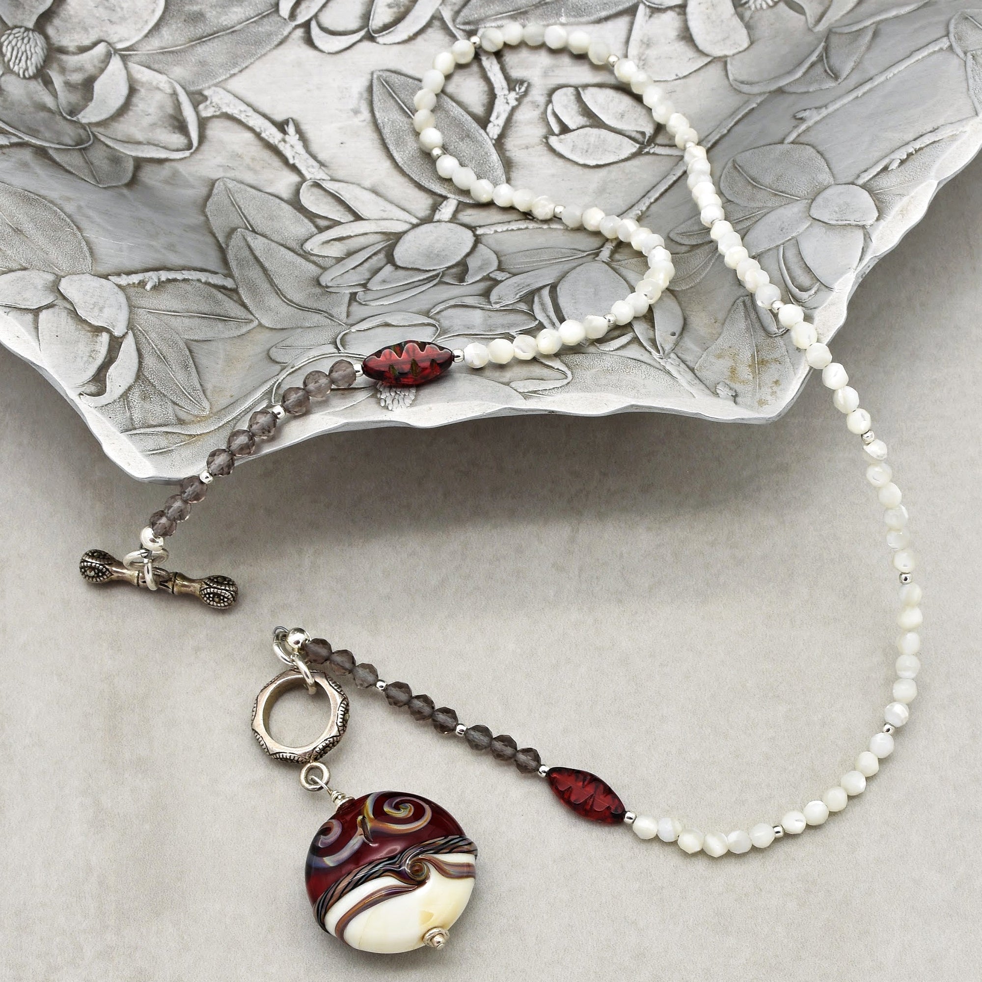 The image shows a necklace. It consists of beads in white, gray, and red colors, and it features a large, decorative round pendant with swirled patterns in shades of red, brown, cream, and white. The necklace also has a metal clasp for fastening.