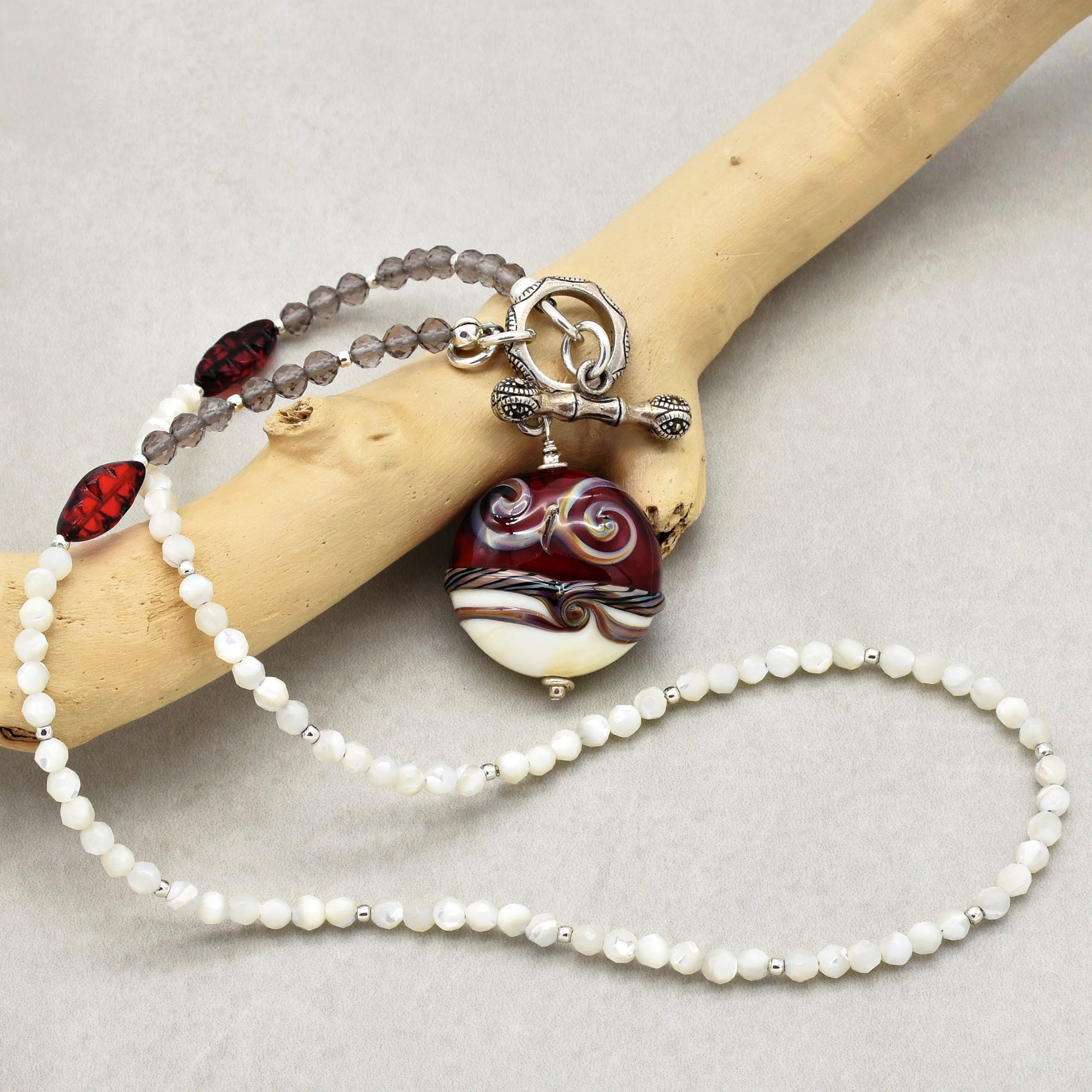 This image shows a necklace. It features a combination of white and translucent beads with accents of larger, decorative beads, and a prominent round pendant with a swirled design in red, white, and black colors. The necklace also has a unique toggle clasp.