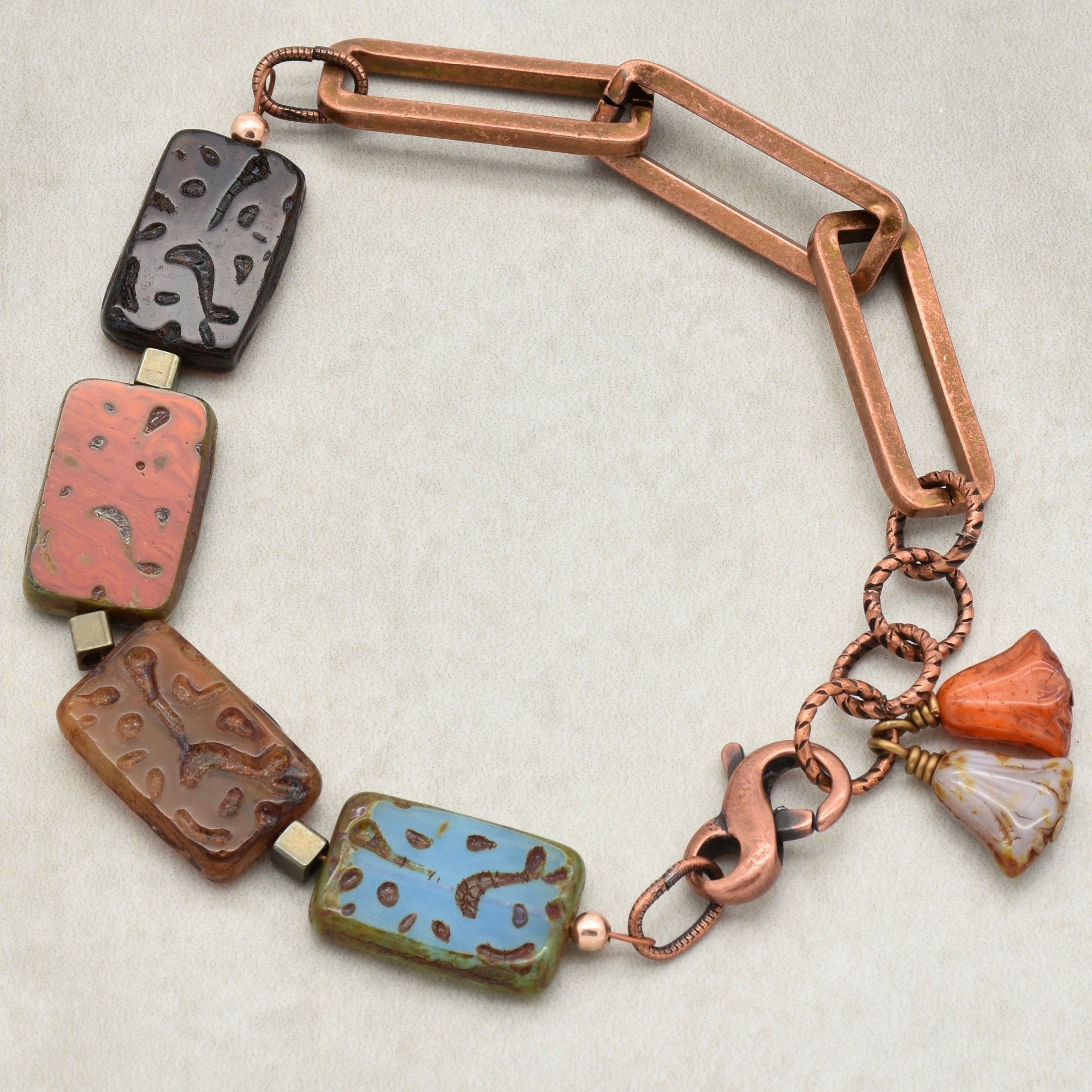 This image shows a bracelet. It features a combination of rectangular beads with intricate patterns and copper chain links, along with a clasp and some small decorative charms. The length and structure indicate it is designed to be worn around the wrist.