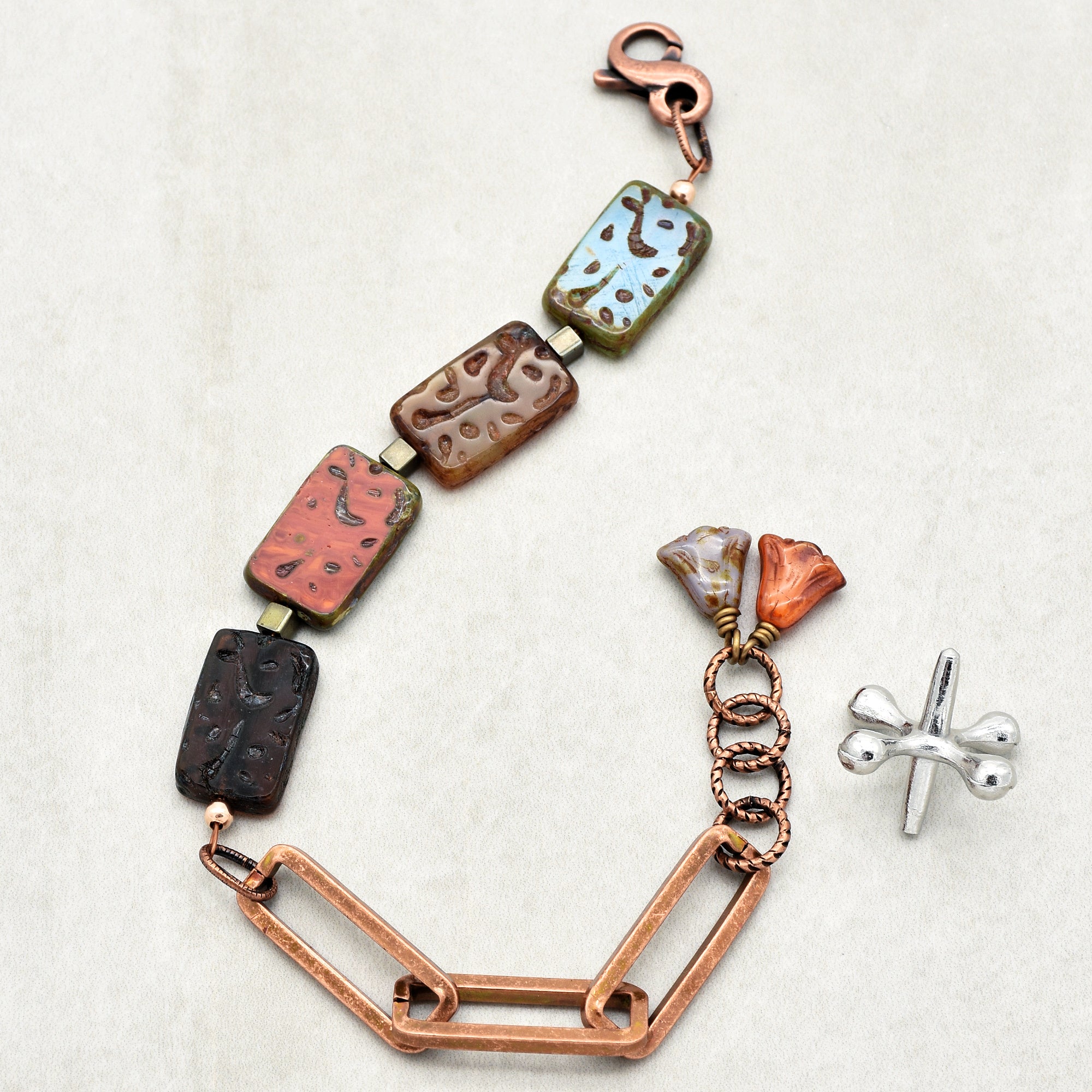 This image shows a bracelet. The jewelry piece features rectangular, multicolored beads, linked by small metallic connectors, and has a clasp at the top for fastening. There is also a decorative element with small flower-shaped beads and rings, along with a separate metallic piece on the side.