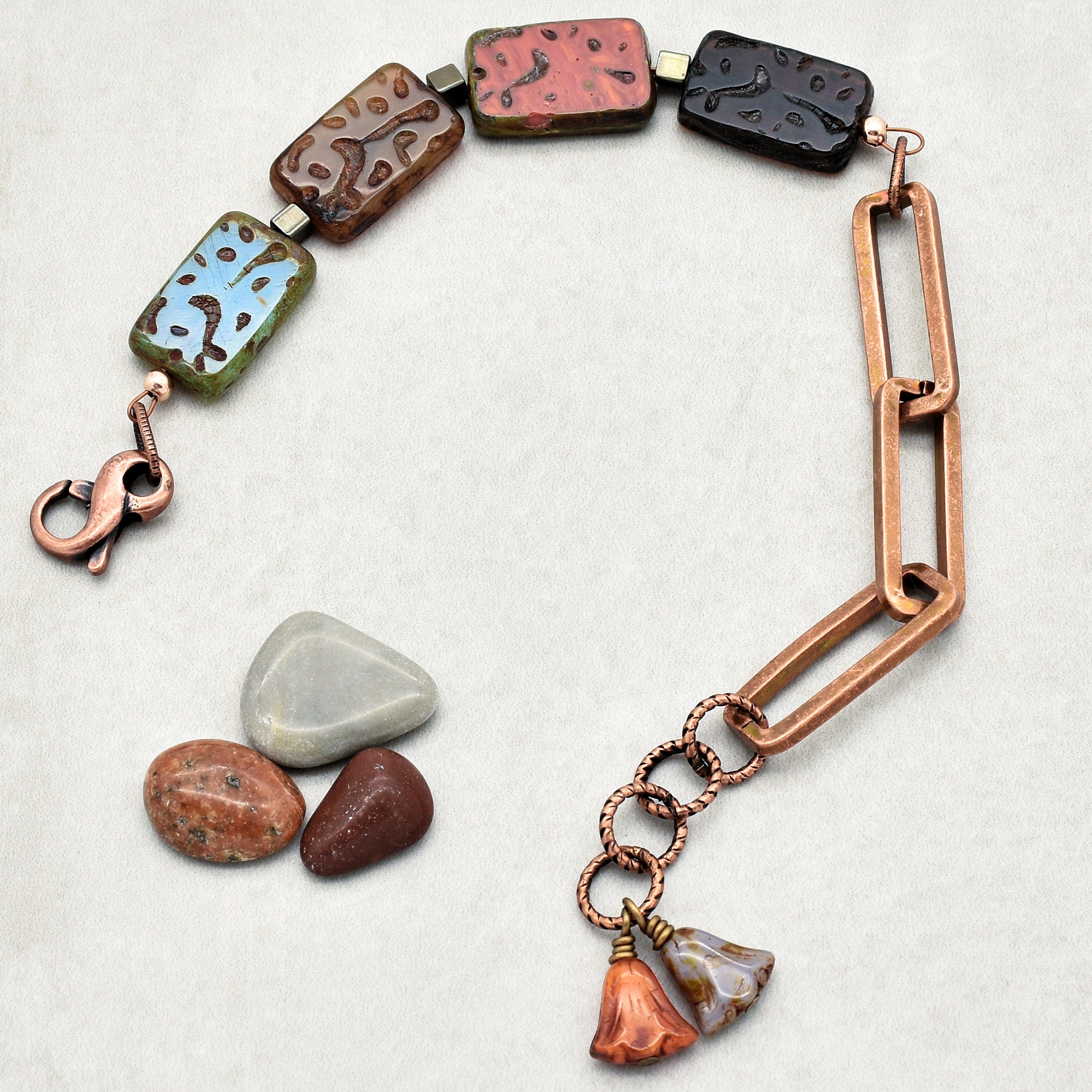 This image shows a piece of jewelry that is a bracelet. The bracelet features a combination of rectangular beads with patterns, large copper links, copper circular links, and two flower-shaped charms hanging from the circular links. The overall design suggests it is meant to be worn around the wrist.