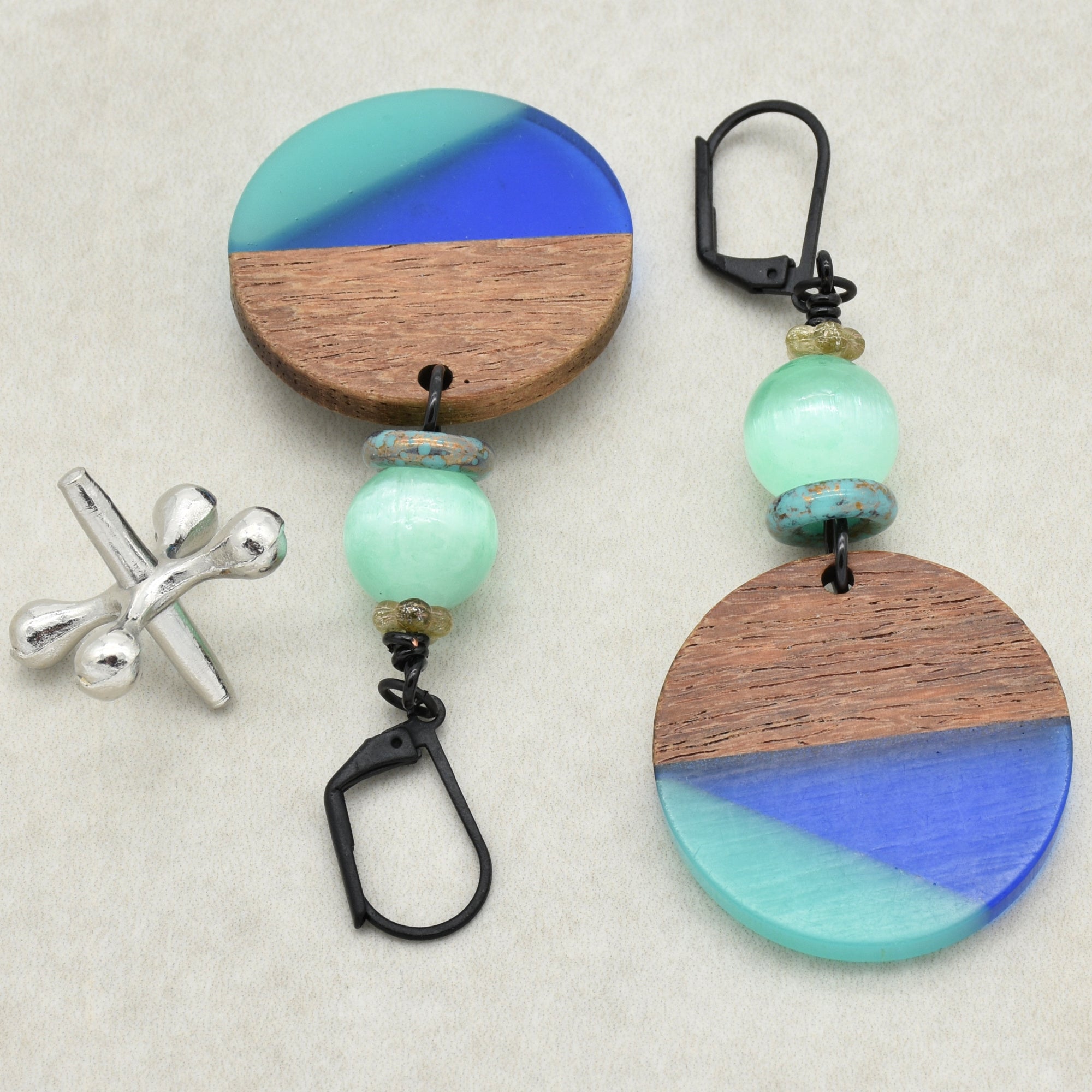 The image shows a pair of earrings. The earrings feature a circular design with a combination of wood and blue-green resin or glass elements, along with additional small beads and a black clasp for fastening.