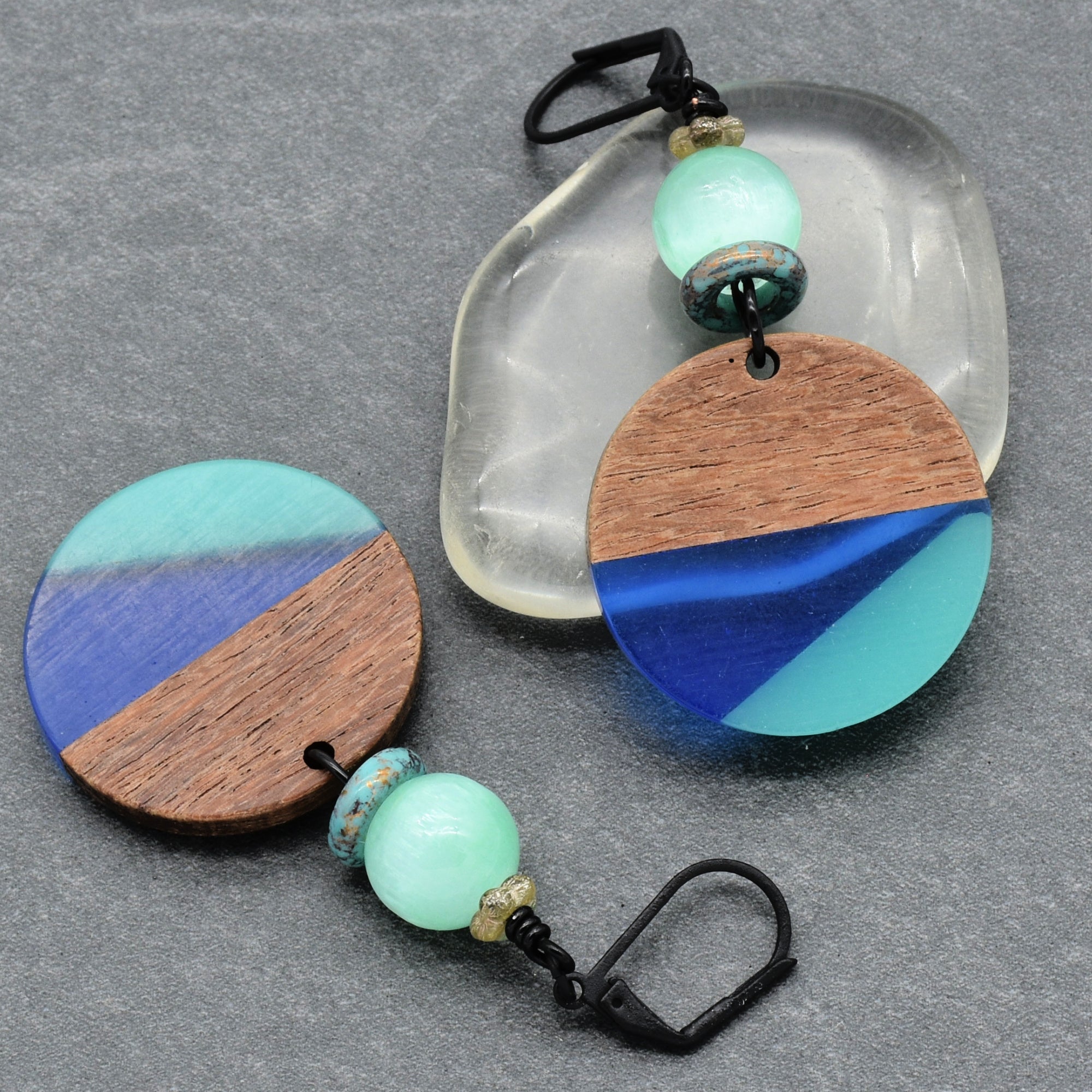 The image shows a pair of earrings. They have circular wooden and resin pieces with blue and turquoise coloring, along with beads near the hooks.