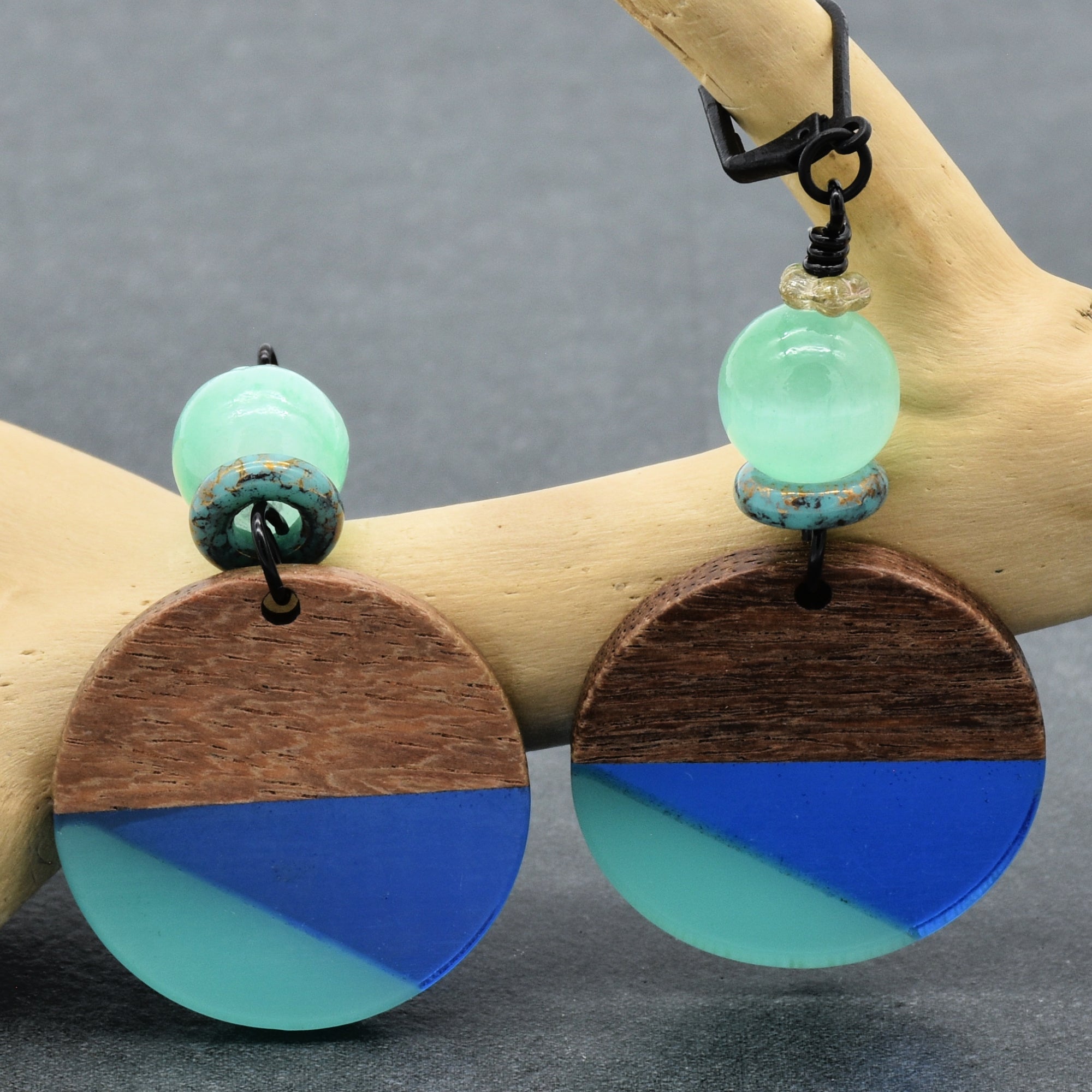 The image shows a pair of earrings. They consist of circular wooden elements with a design divided into two colors (blue and green) and have beads above the wooden circles. The earrings are hanging from a display.