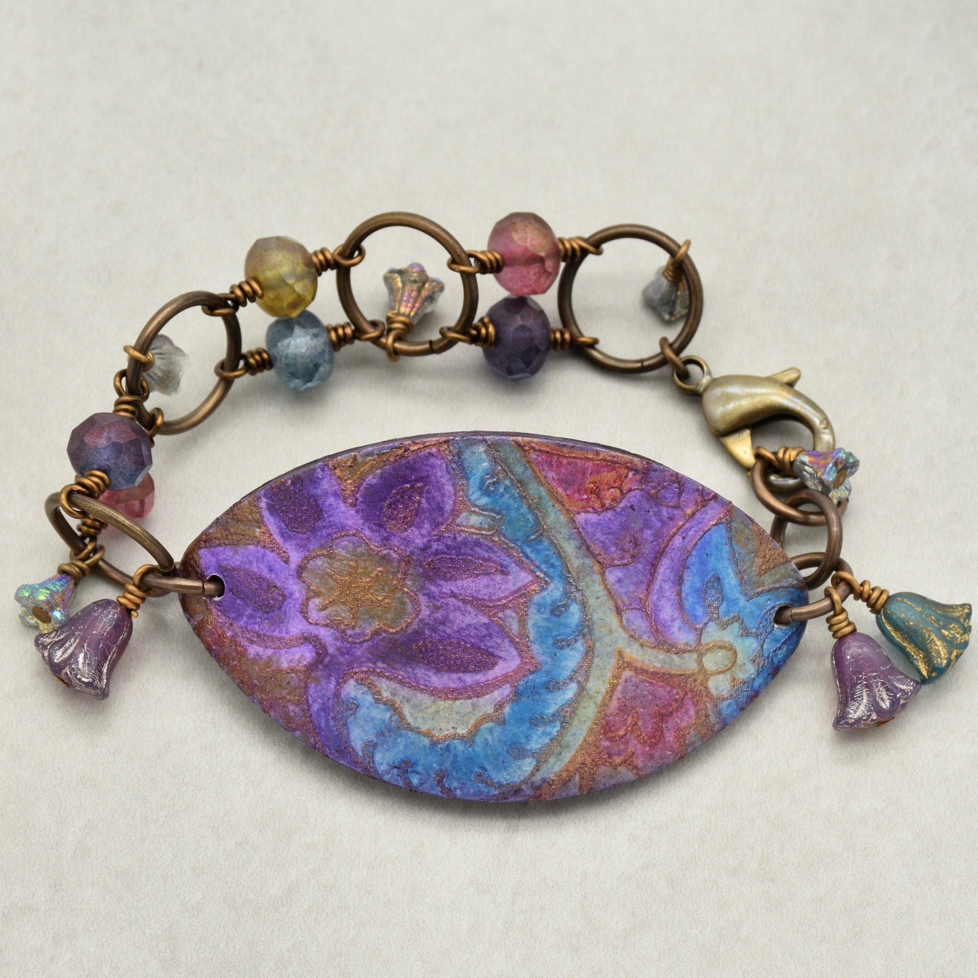 This image shows a bracelet. It features a central decorative piece with a purple and blue floral pattern, connected to a chain of circular links adorned with colorful beads. The clasp on the right side indicates that it is a bracelet.