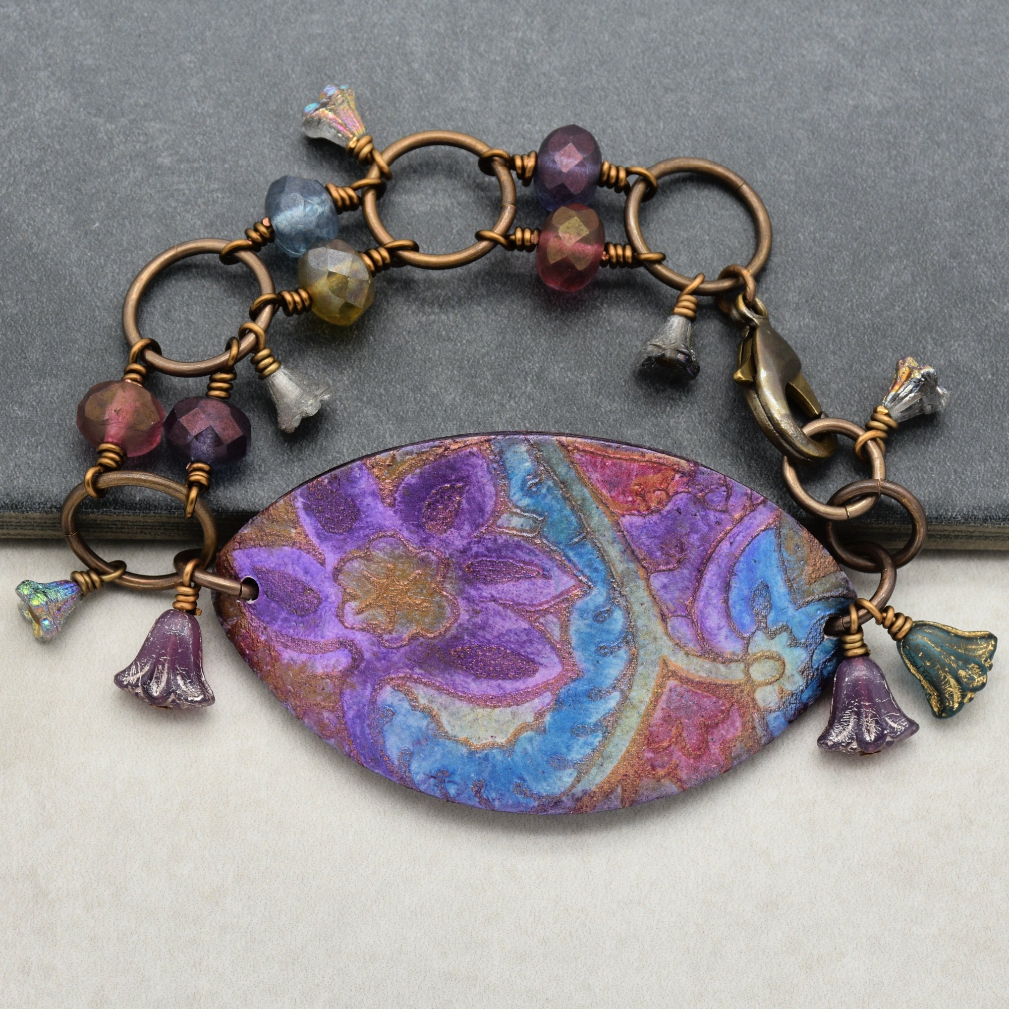 This image shows a bracelet. The jewelry piece is circular and has a clasp, which is characteristic of a bracelet. It features a central decorative element with colorful floral patterns and is adorned with beads and small flower-shaped charms around the chain.