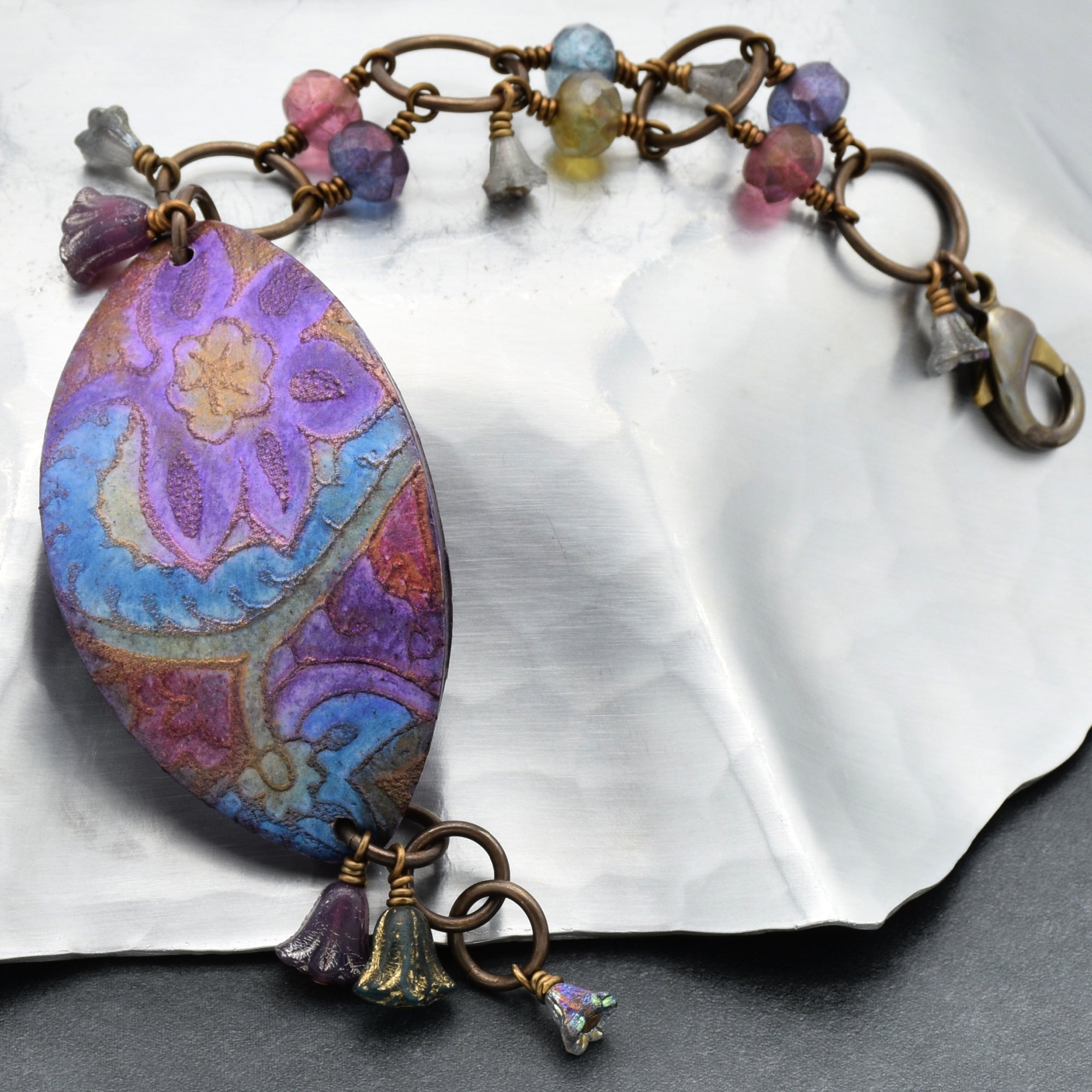 The image shows a bracelet. It features a decorative, elongated central piece with a colorful floral pattern in purple, blue, and gold tones. The bracelet also has various small beads and charms attached to a chain with circular links and a lobster clasp.