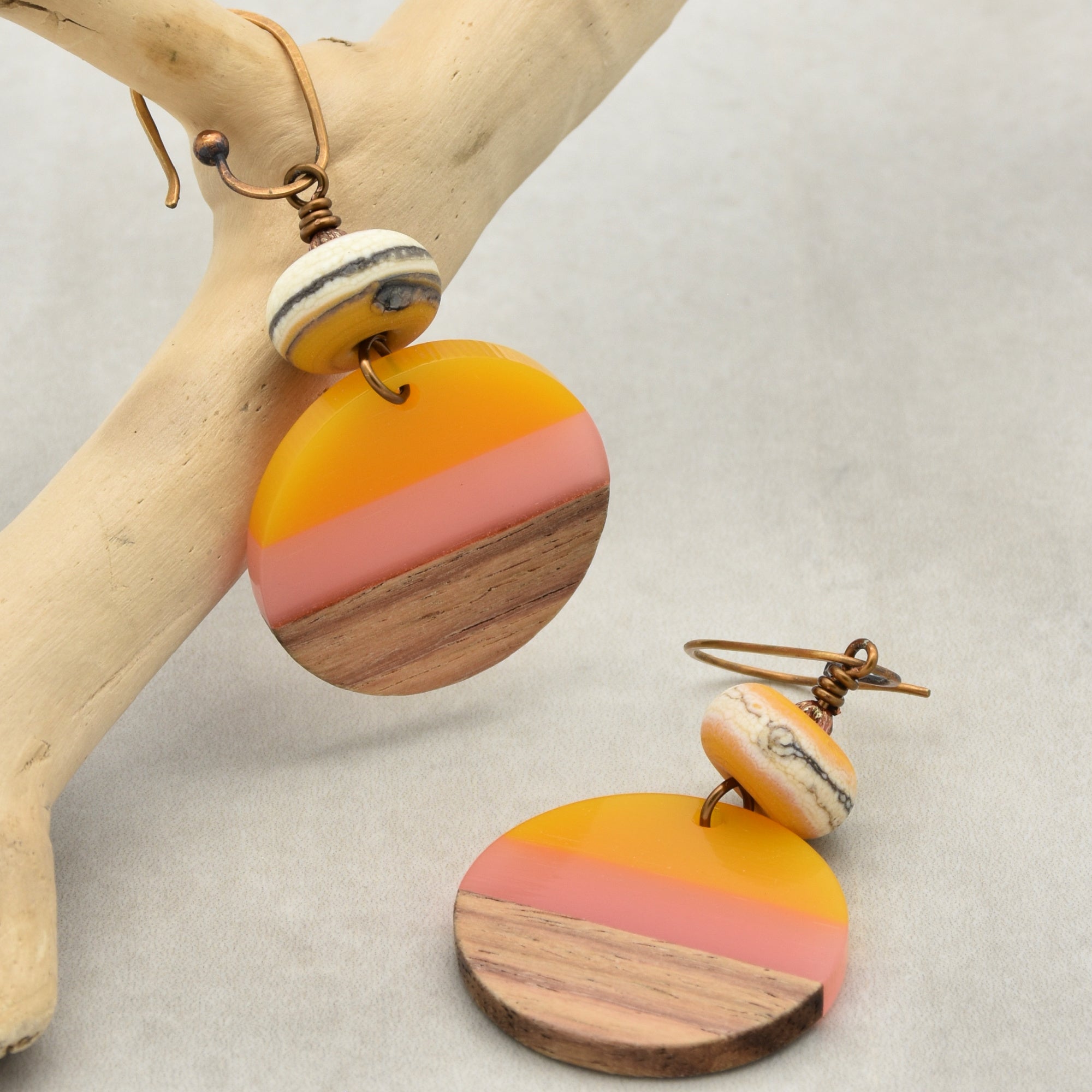 The image shows a pair of earrings. They have a circular design with a combination of wood and colored materials in yellow and pink tones, and are attached to metal hooks.