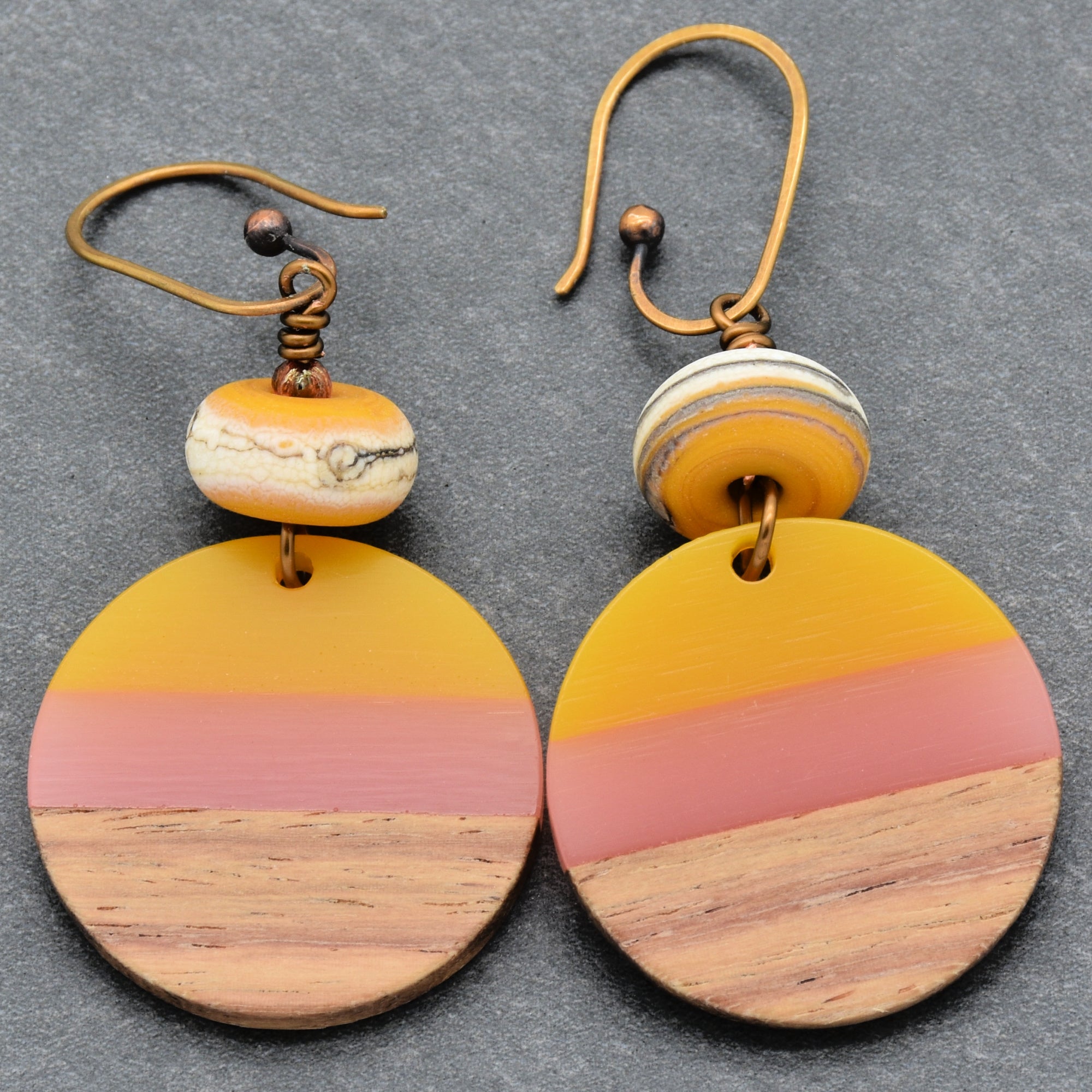 The image shows a pair of earrings. They are circular and have a design with three distinct color sections: yellow on the top, pink in the middle, and a wood-textured section on the bottom. The earrings also have a small round bead above the circular piece and have hooks for wearing.