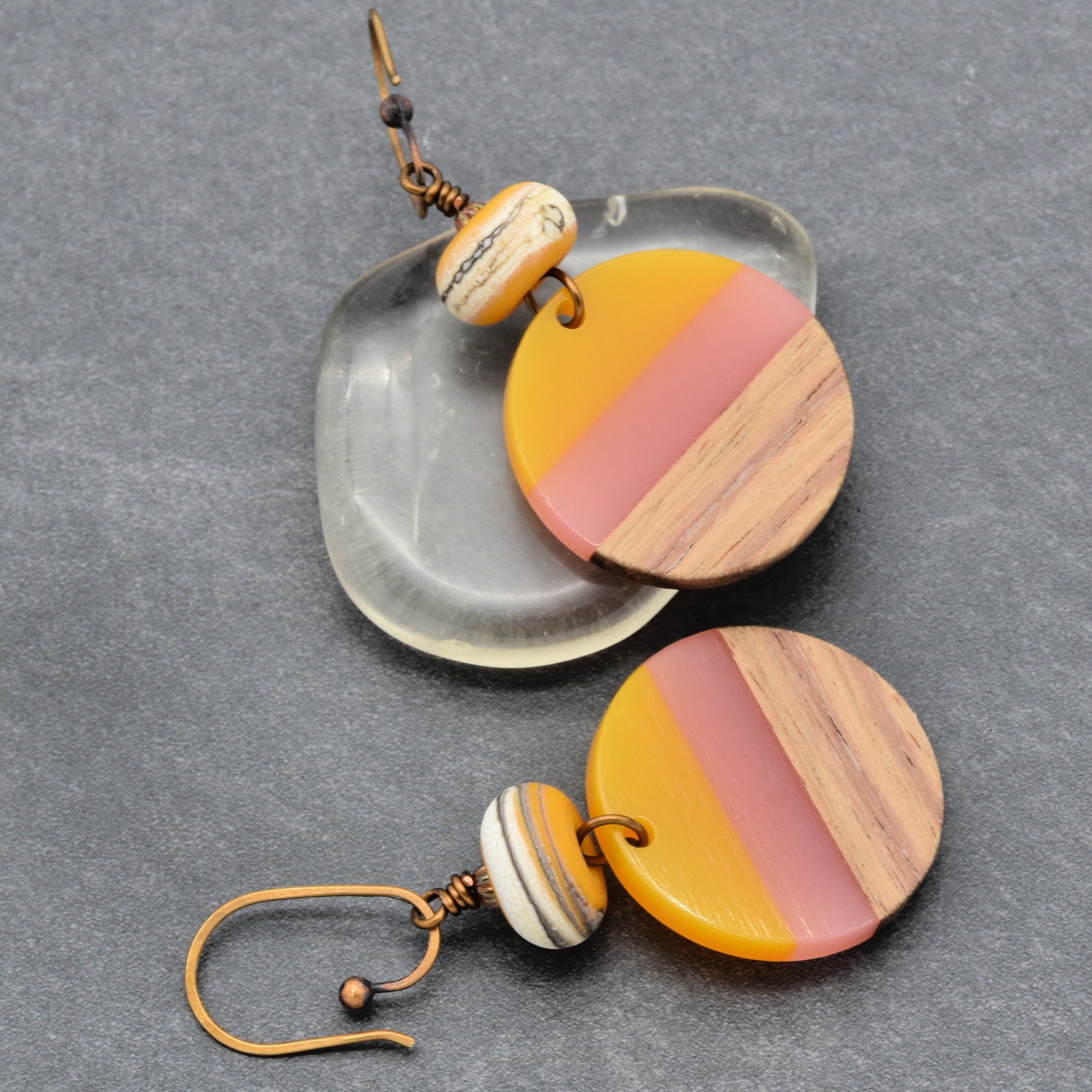 The image shows a pair of earrings. They have a round, multicolored design featuring yellow, pink, and wood-like sections, along with a smaller bead above the round piece, attached to ear hooks.