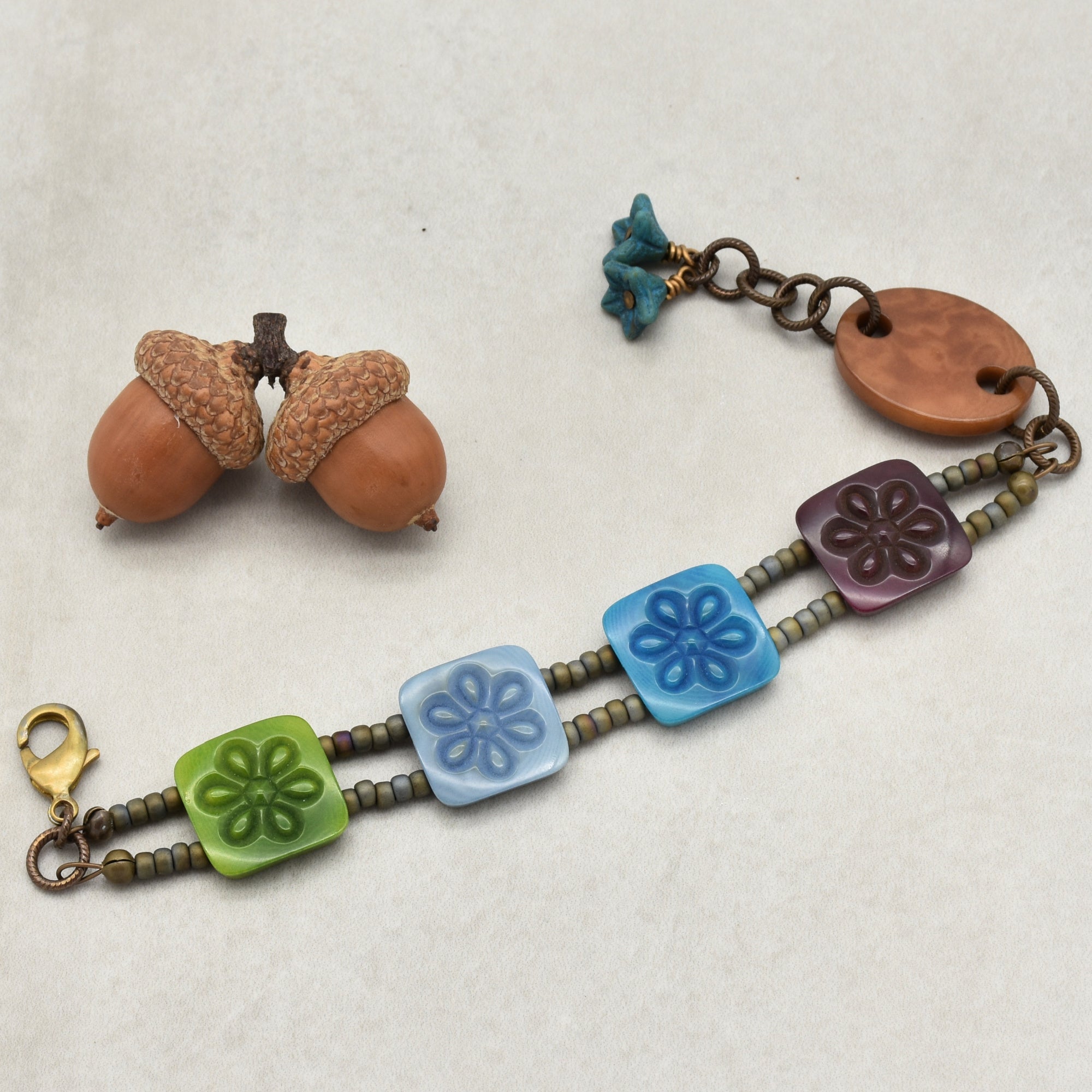 This image shows a bracelet. The bracelet has two strands with beads and square floral-patterned pieces in green, various shades of blue, and purple, connected by a metal clasp and chain.