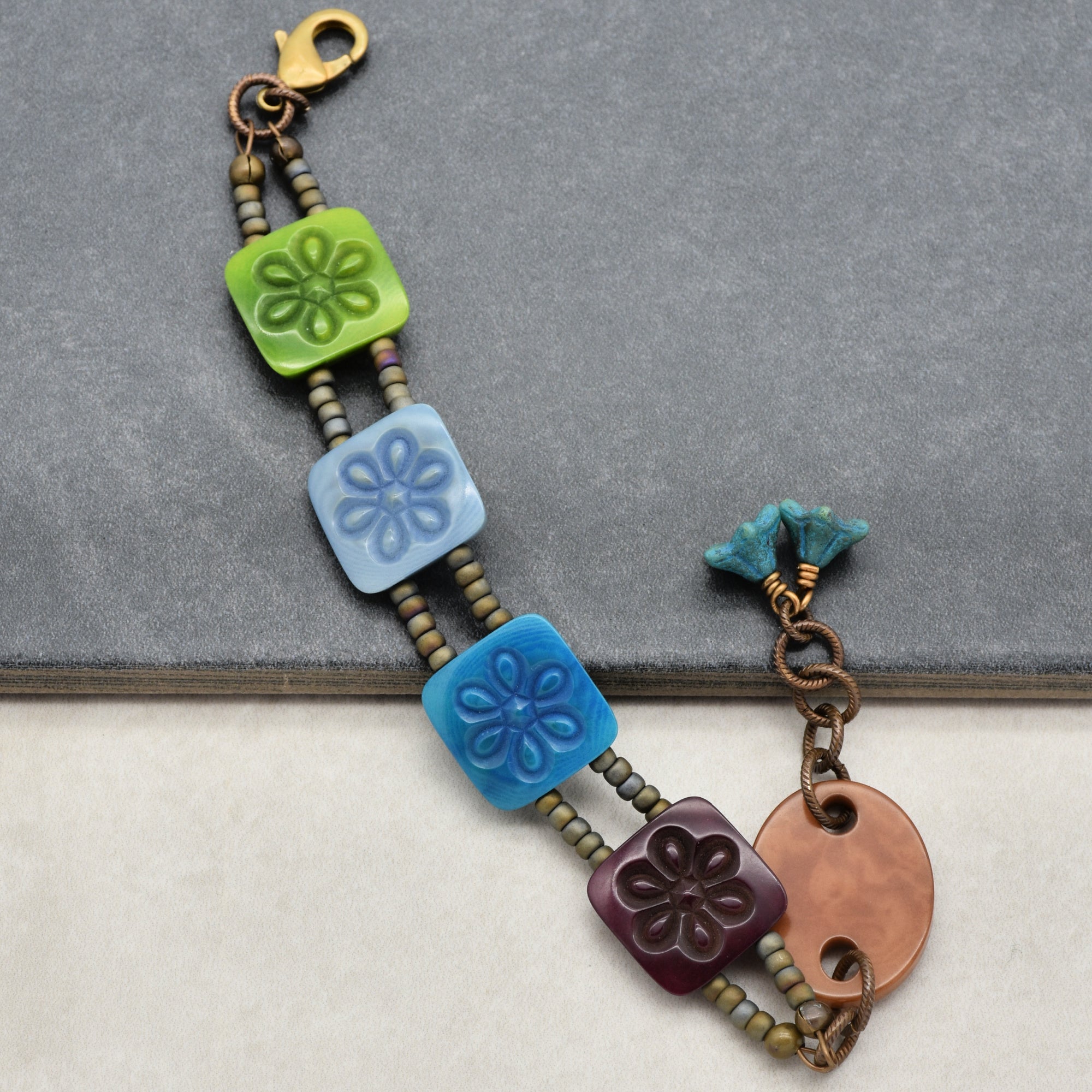 The image shows a bracelet featuring three square tiles with floral designs in green, light blue, and dark blue. It is strung with small round beads and includes a round textured copper piece and turquoise-colored accents near the clasp.