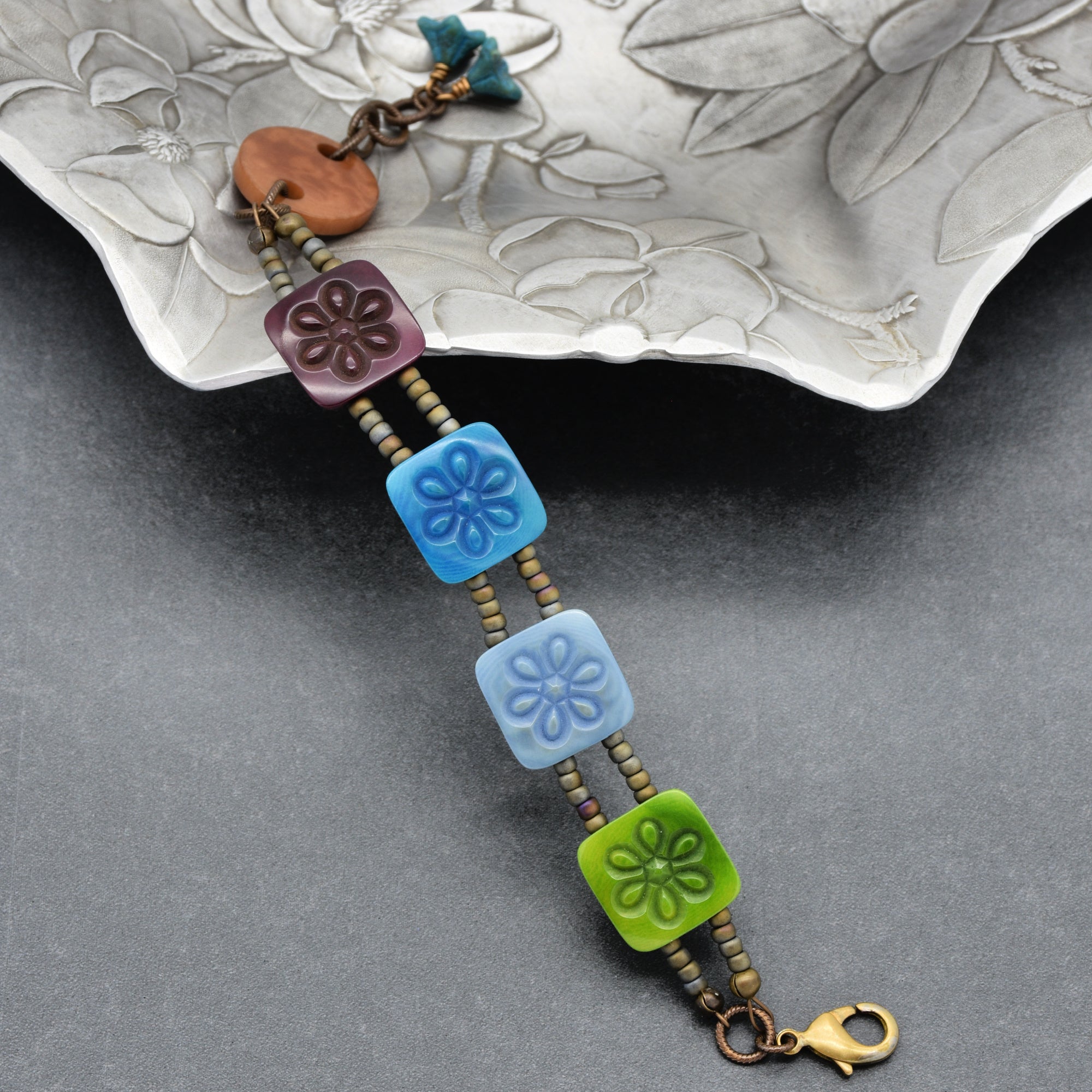 This image shows a bracelet. It features a design with square beads in blue and green colors, each with a floral pattern, along with smaller round beads in between. The bracelet has a clasp closure at one end.