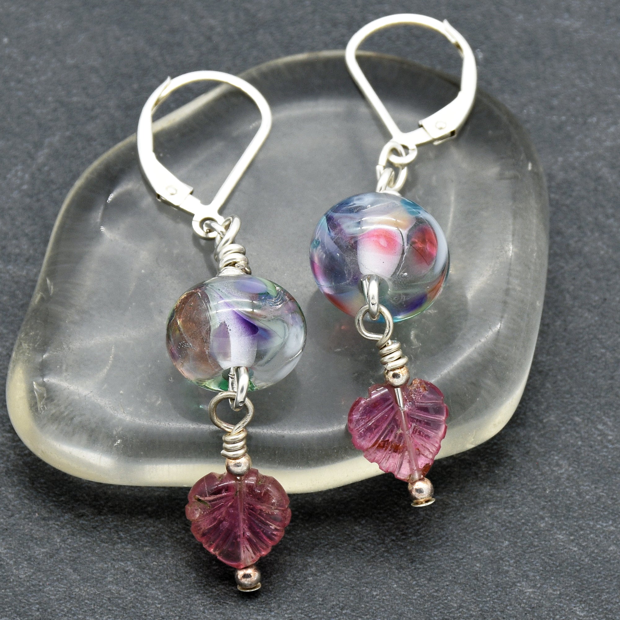 The image shows a pair of earrings. They have hooks at the top for attaching to pierced ears, and each earring features a round, colorful bead with a small purple leaf-shaped charm dangling below it.