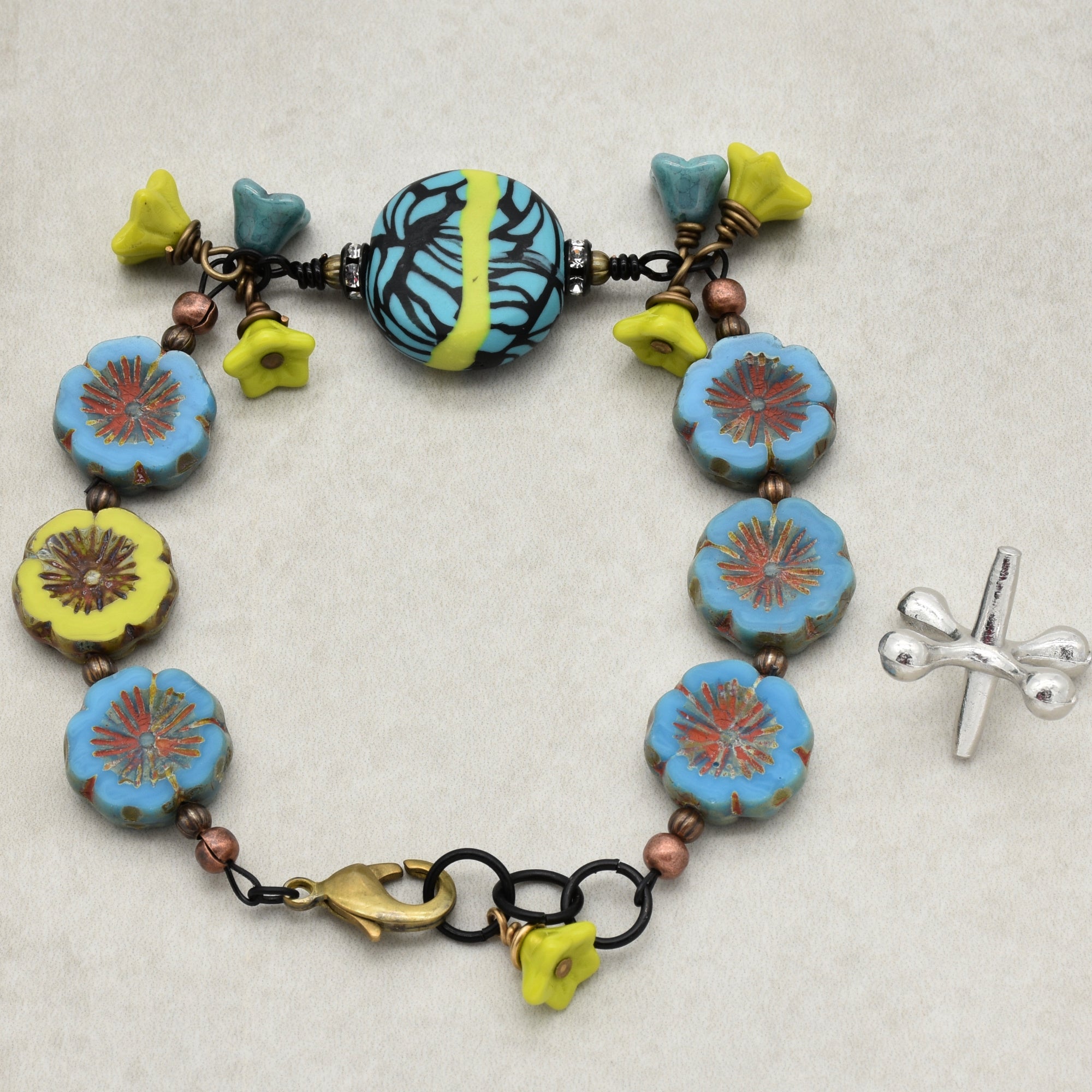 This image shows a bracelet. The piece features flower-shaped beads in blue and yellow, along with a larger patterned bead and various smaller decorative elements. The size, clasp, and design indicate that it is a bracelet rather than a necklace or earring.