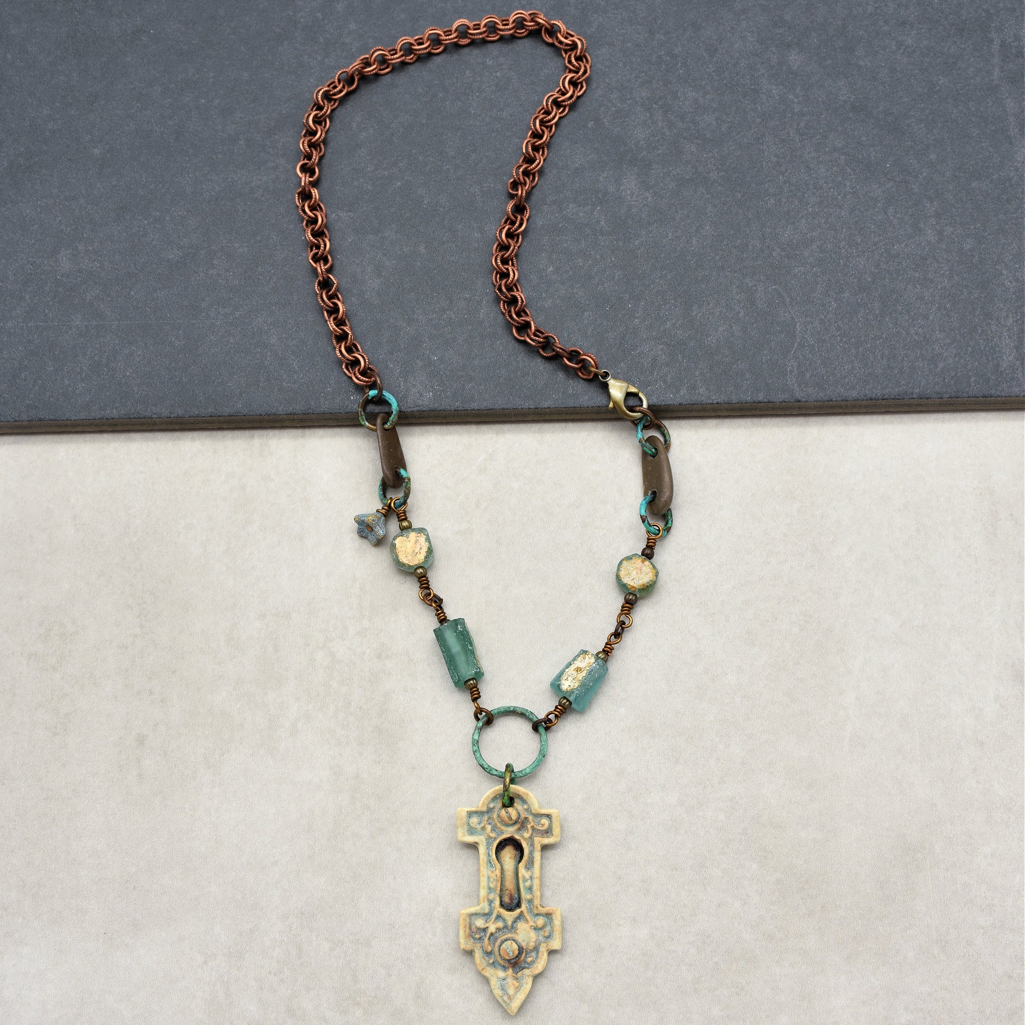 The image shows a necklace. It features a chain with copper-colored links and several turquoise beads, with a decorative pendant resembling an ornate keyhole at the center.