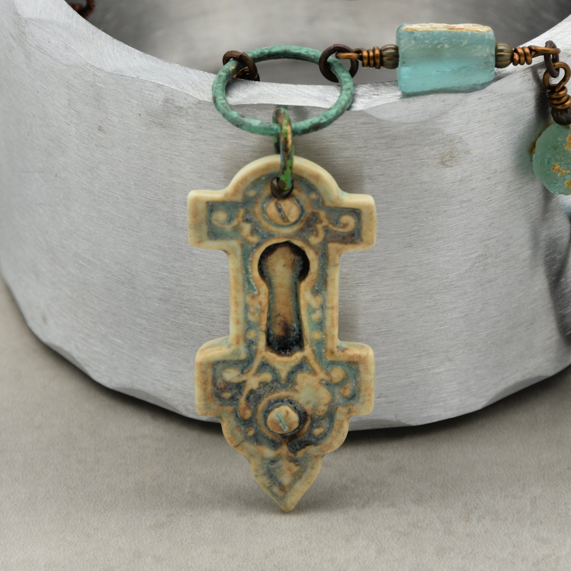 The image shows a piece of jewelry that is a necklace. The necklace features a keyhole-shaped pendant with an ornate design and a weathered look, hanging from a greenish ring that is part of the necklace chain. The chain also has several beads.