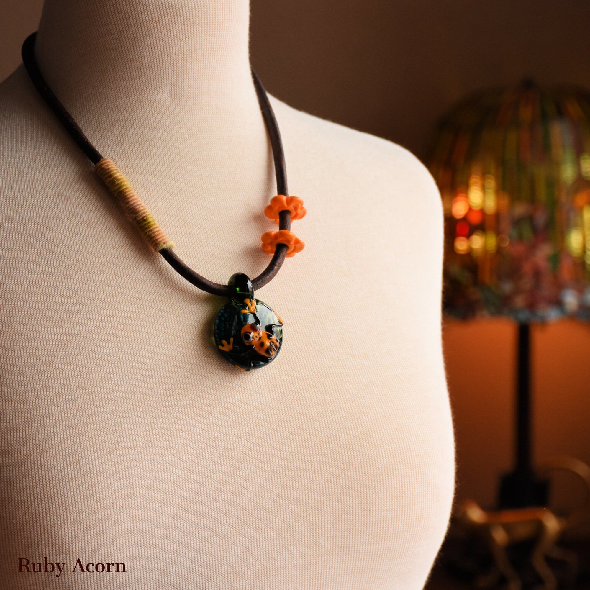 This image shows a necklace. It features a cord with decorative elements, including an orange frog charm on a small circular pendant, and two orange flower-shaped beads. The necklace is displayed on a mannequin.