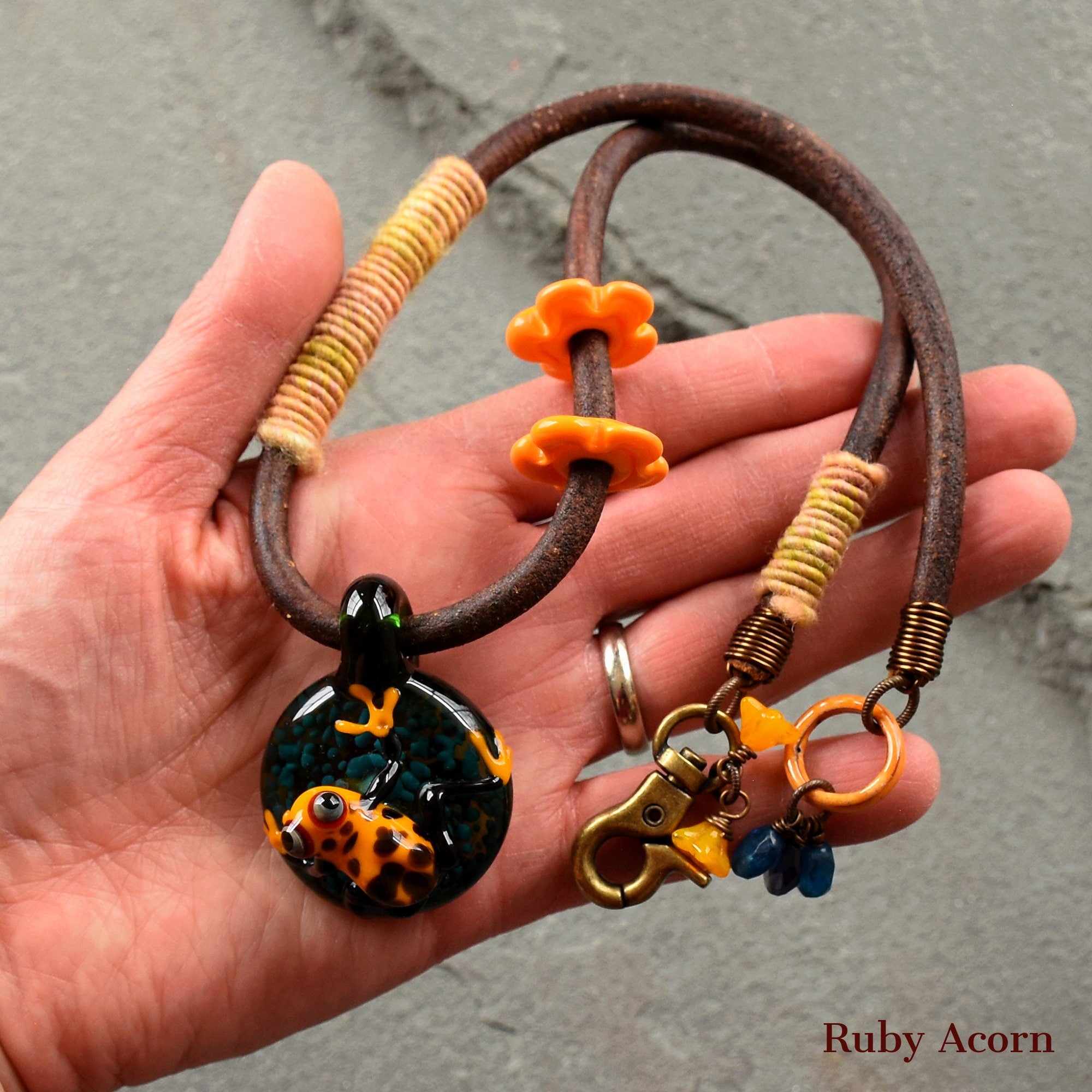 This is a necklace. The image shows a pendant hanging from a leather or cord necklace, which is indicated by the clasp and the length of the material. The pendant features a decorative design with what looks like a small frog or similar creature. The presence of a clasp further confirms that this item is intended to be worn around the neck.