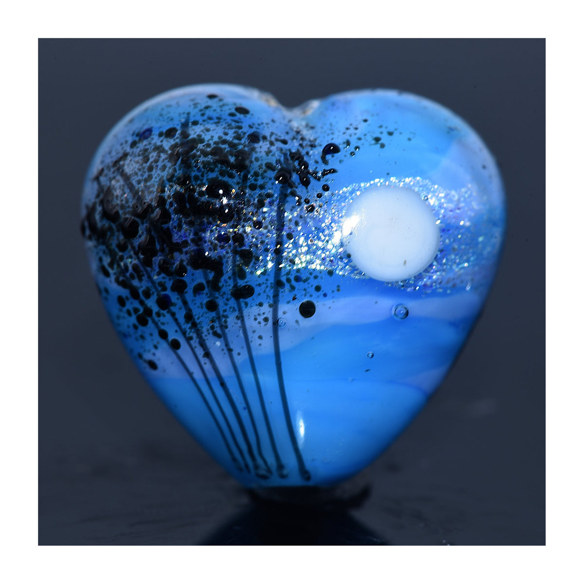 Blue heart-shaped pendant with black speckles on a dark background - Grace Lampwork Beads available at Ruby Acorn.