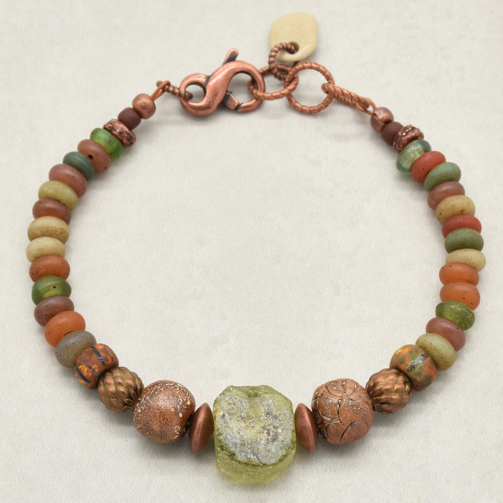 A handmade beaded bracelet featuring a mix of earthy Indonesian glass beads in terracotta, olive green, ochre, and brown, accented by textured copper beads, a central raw green ancient Roman glass focal, artisan ceramic beads, and a copper clasp on a light beige background - made with love in Colorado by Ruby Acorn. 